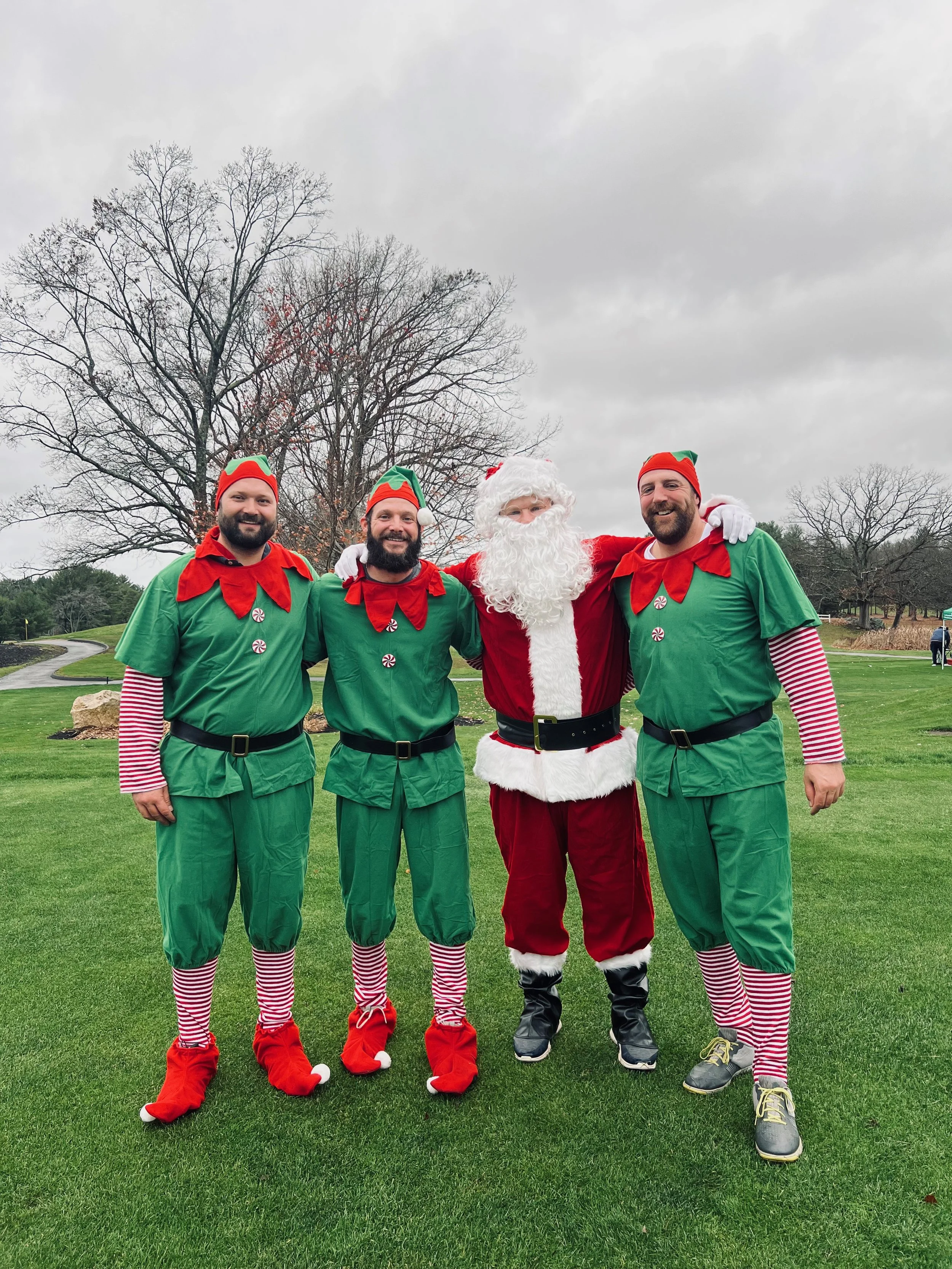 Santa Claus and three men dressed as Christmas elves pose on a grassy golf course.