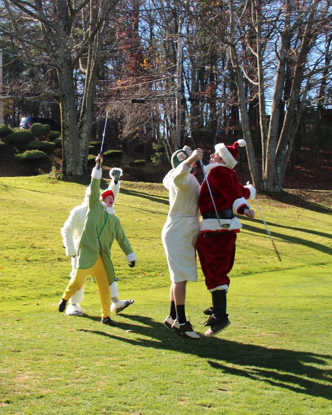 Santa Claus and two people in festive costumes play golf on a sunny lawn.