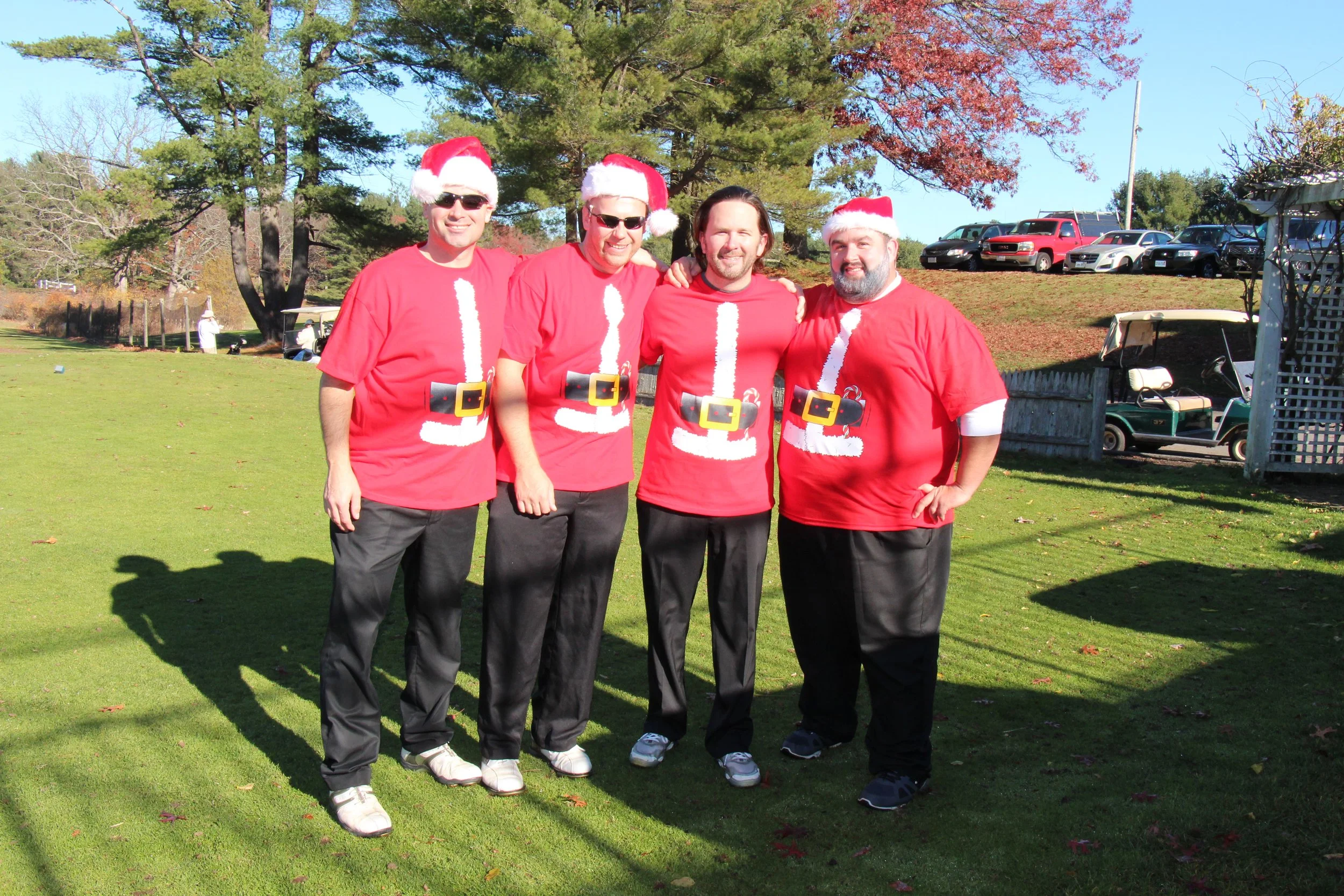 Four men posing outdoors in matching Santa Claus T-shirts and Santa hats on a sunny golf course.