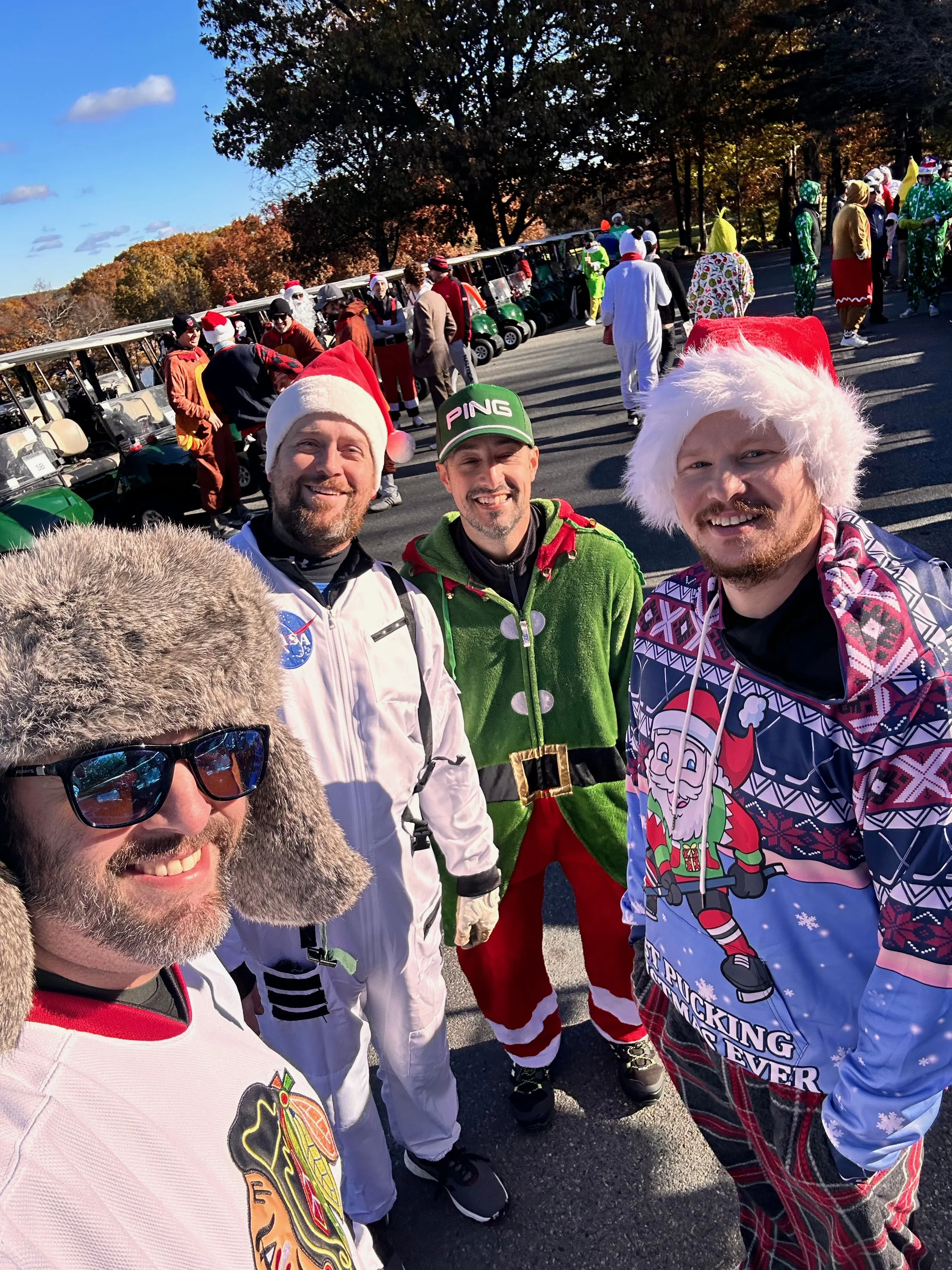Large group in colorful Christmas costumes posing for a selfie beside golf carts on a sunny autumn day