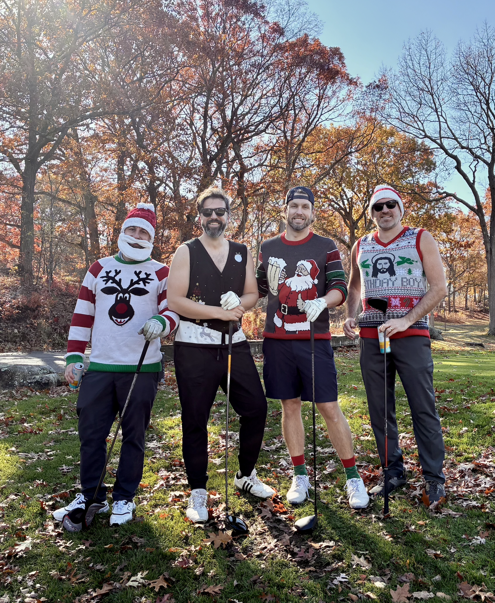 Four men in festive Christmas sweaters and hats pose with golf clubs on a sunny autumn golf course.