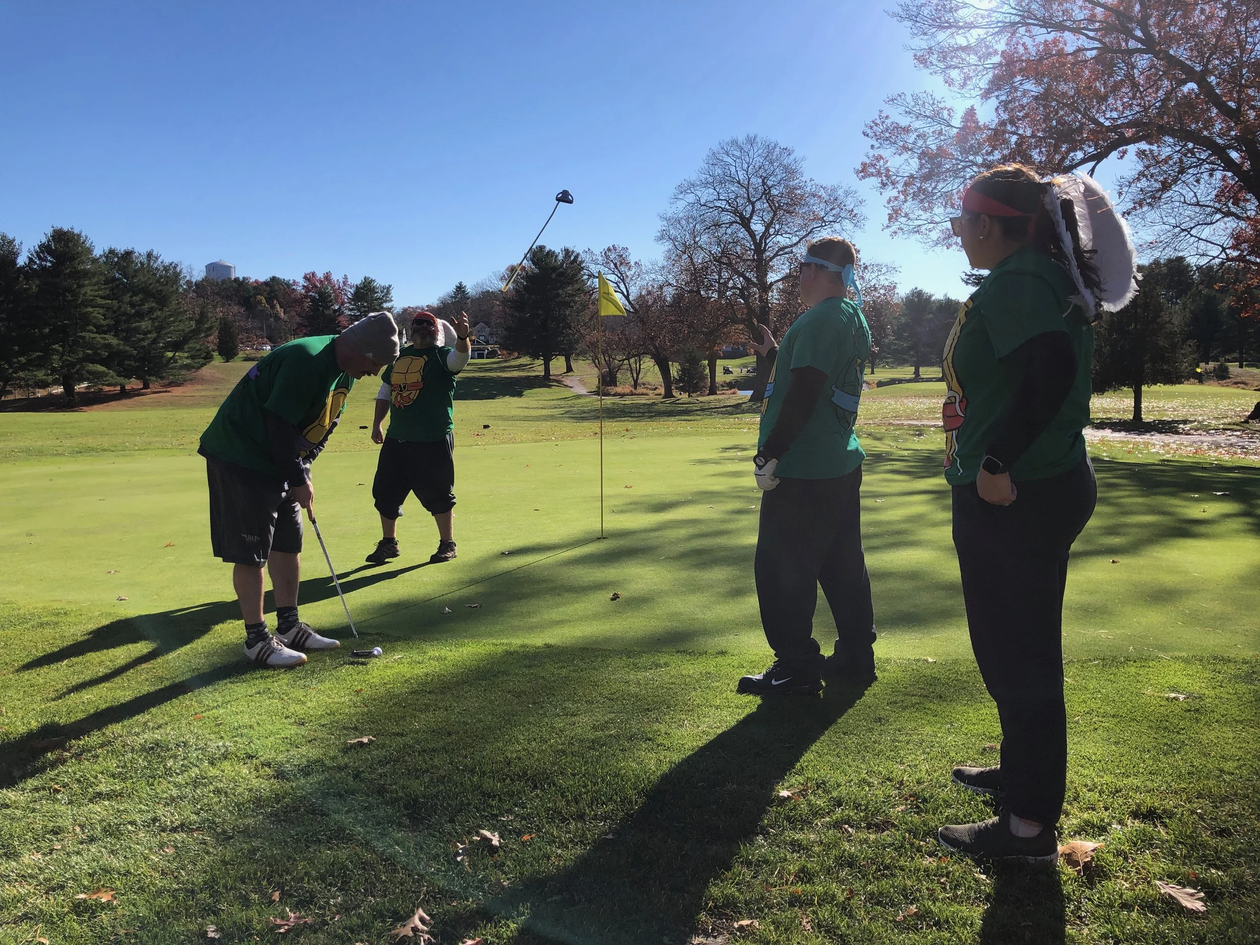 Group of golfers in green shirts playing on a golf course beside a putting green and flagstick on a sunny autumn day