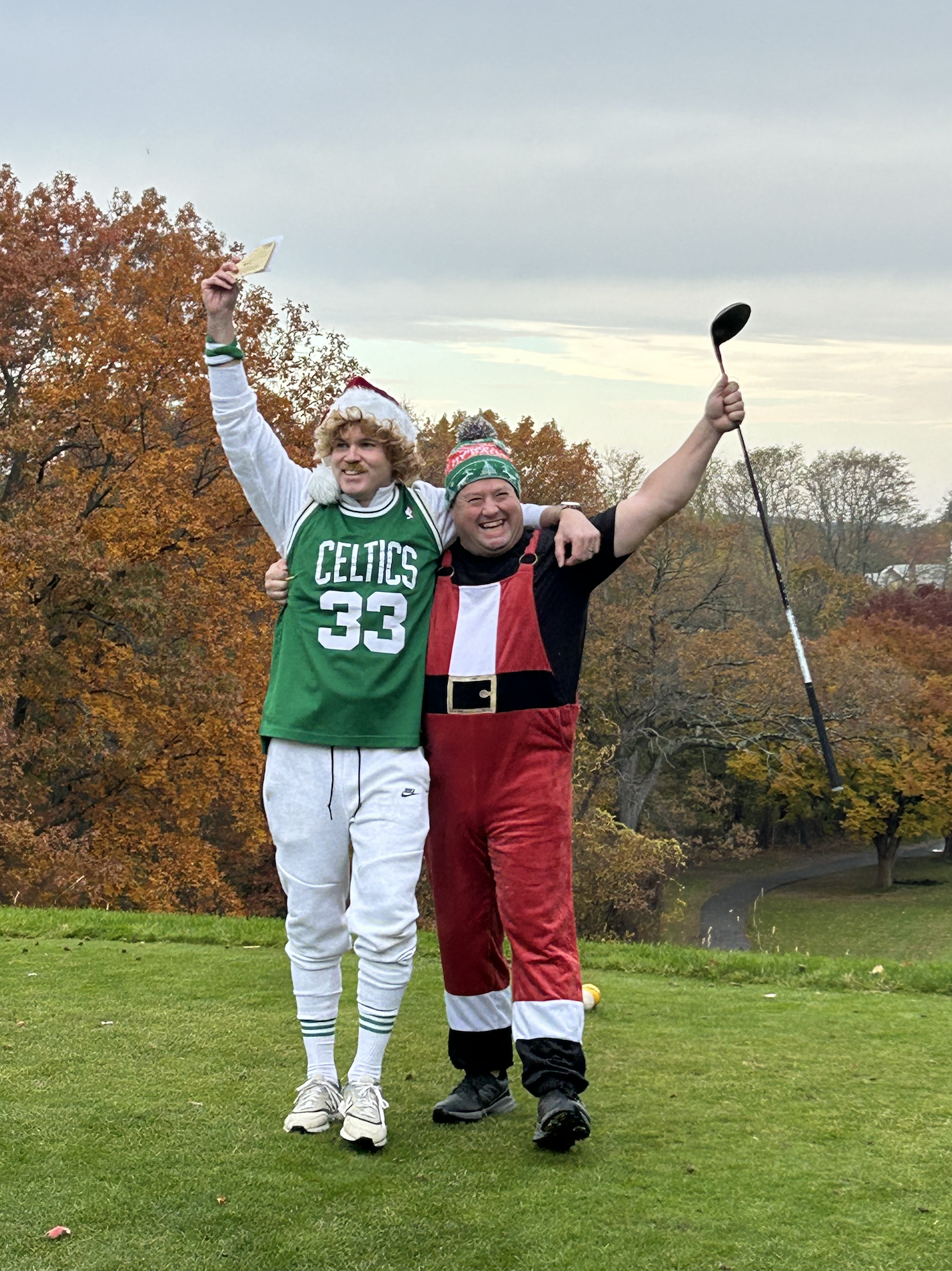 Two smiling men in Celtics and Santa costumes celebrate outdoors on an autumn lawn, one holding a paper and the other raising a golf club.