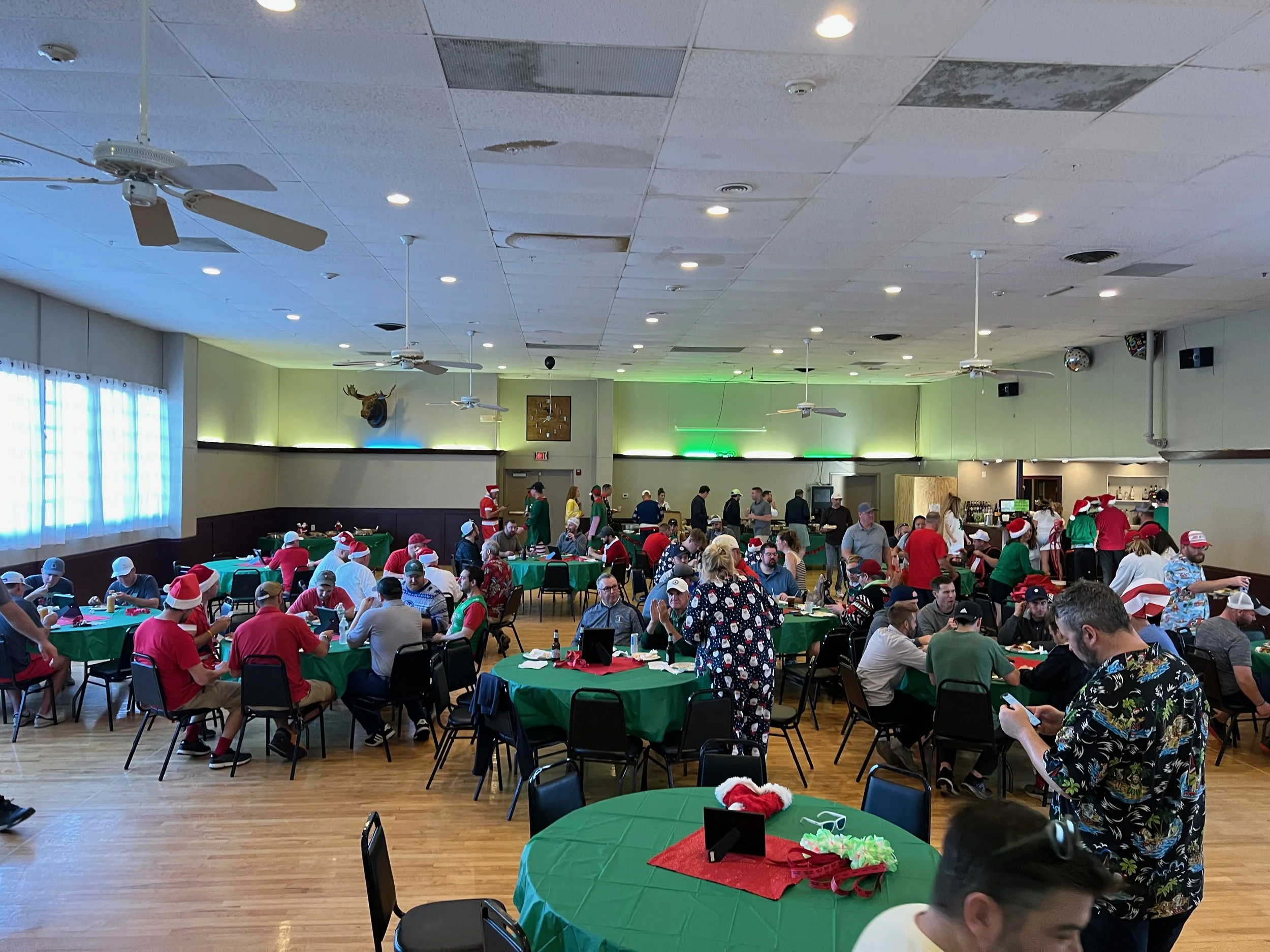 Crowded holiday party in a banquet hall with guests wearing Santa hats, seated at green-covered tables.