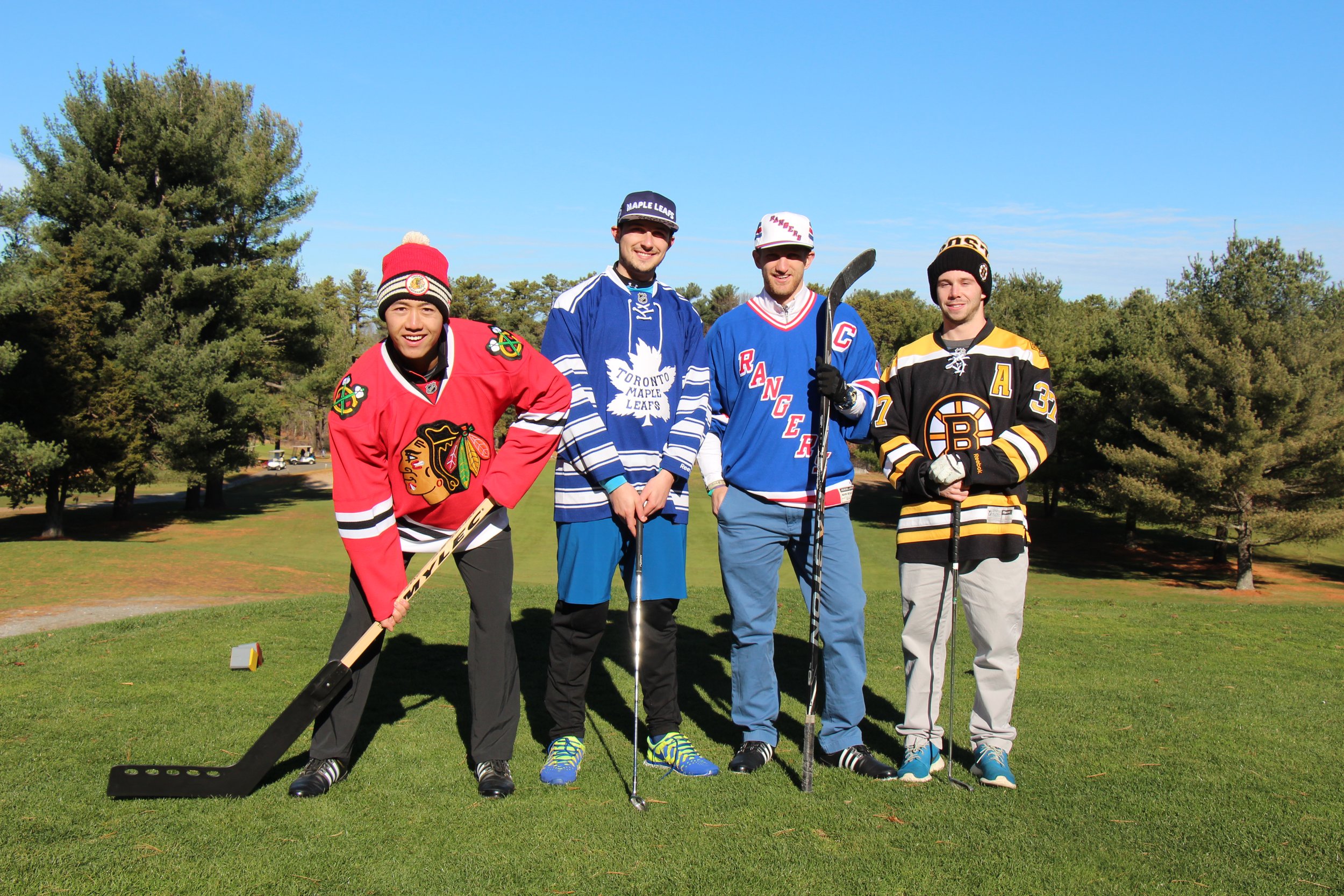 Four men in Chicago Blackhawks, Toronto Maple Leafs, New York Rangers, and Boston Bruins hockey jerseys pose with sticks on a sunny grassy field.