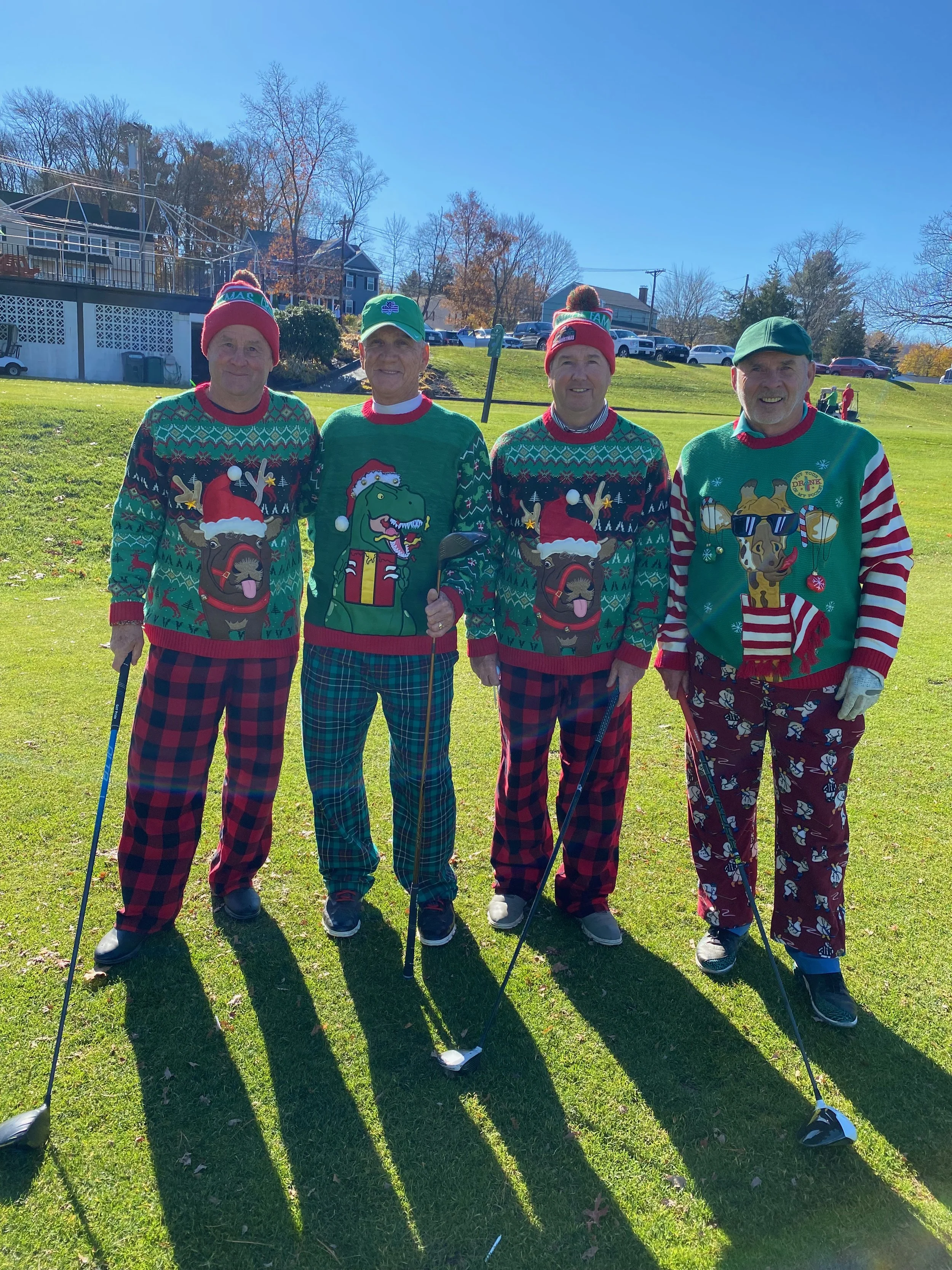 Four men wearing festive Christmas sweaters and hats pose with golf clubs on a sunny lawn.