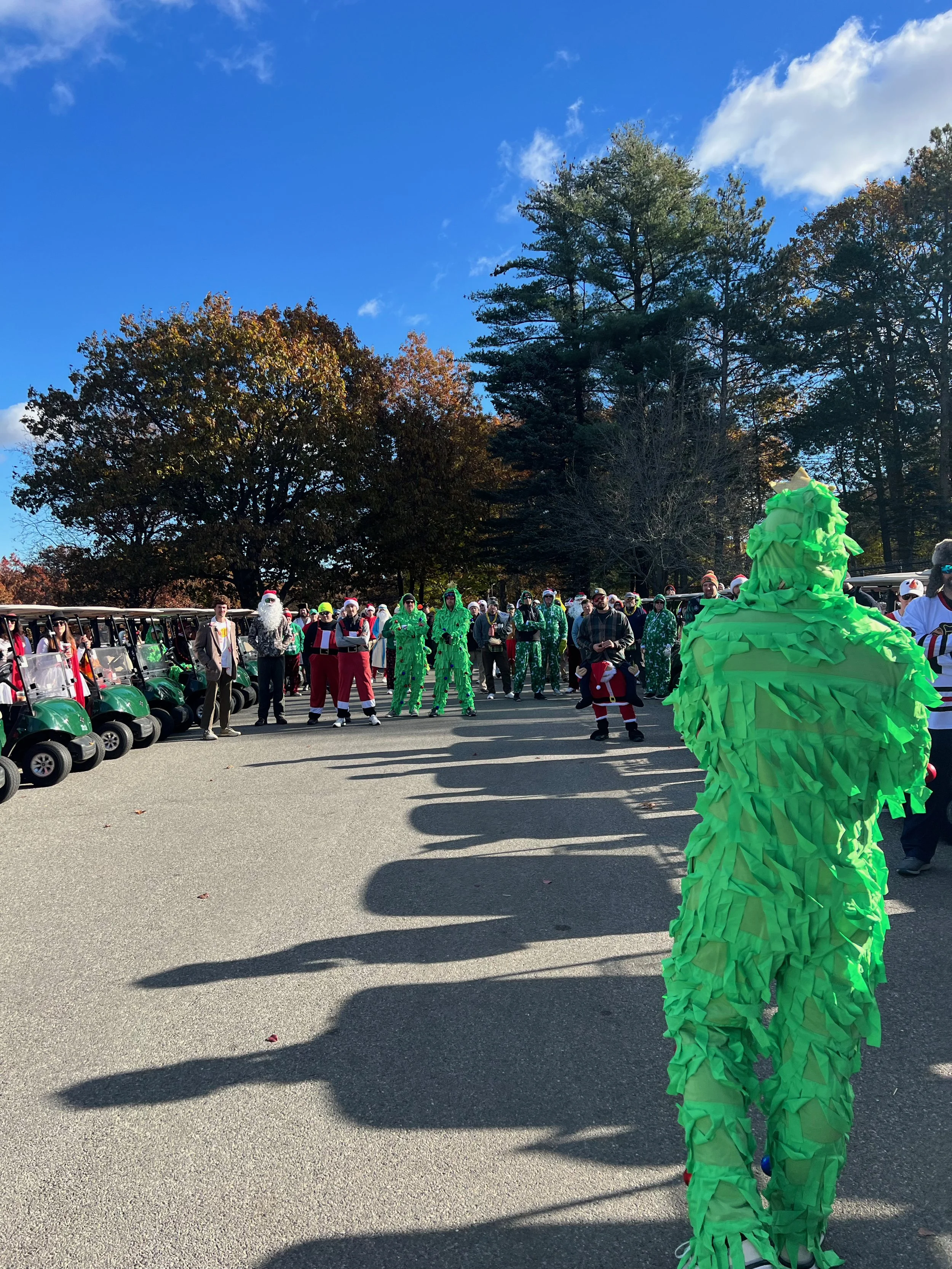 Crowd in Santa and Christmas tree costumes gathered outdoors beside golf carts under a sunny blue sky.