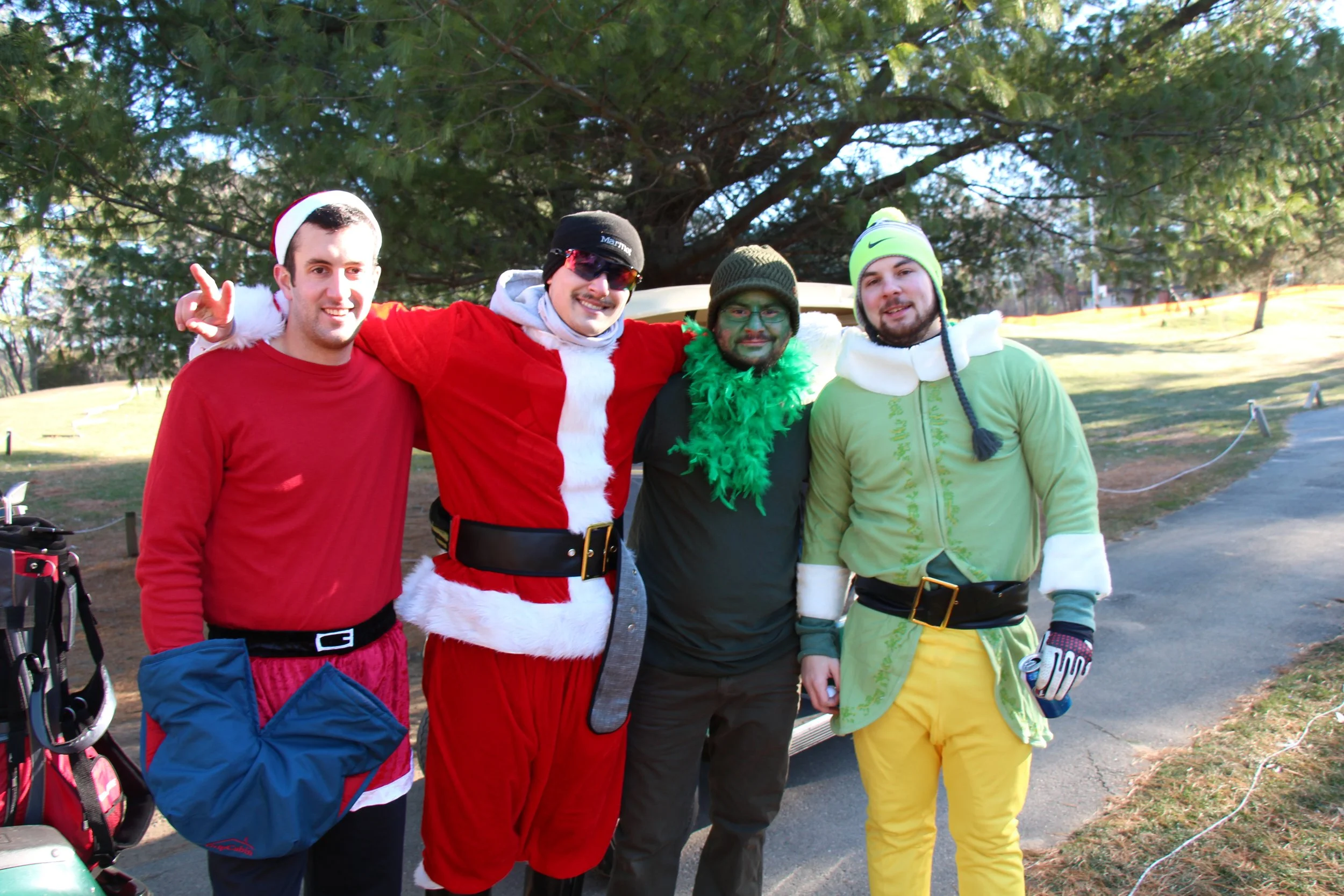 Four men in festive costumes pose outdoors, including a Santa outfit, elf costume, and green feather boa.