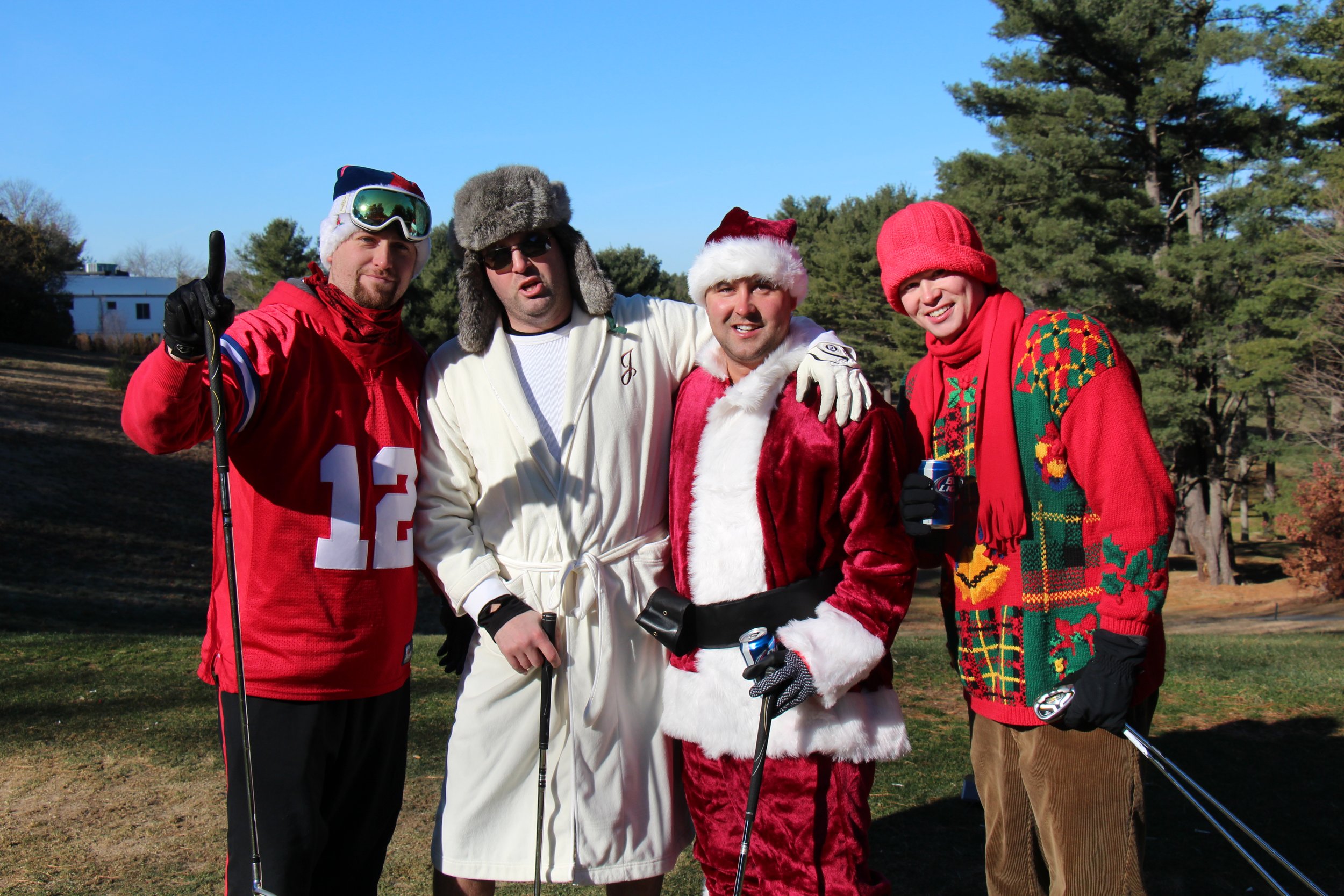 Four men dressed in festive costumes pose outdoors with golf clubs, including Santa Claus, a hockey player, and a man in a Christmas sweater.