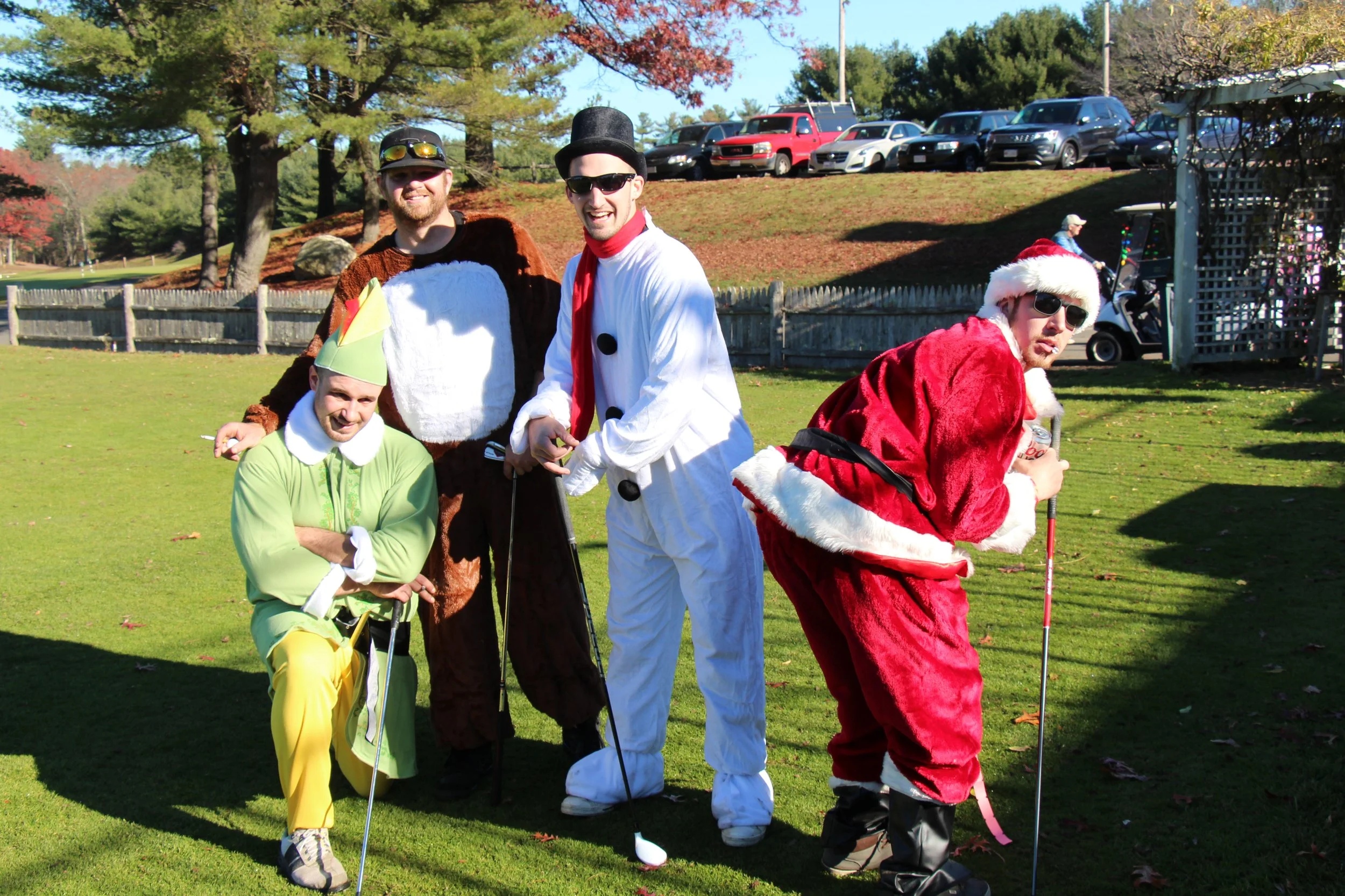 Four adults in colorful costumes, including a snowman and Santa Claus, pose with golf clubs on a sunny golf course.