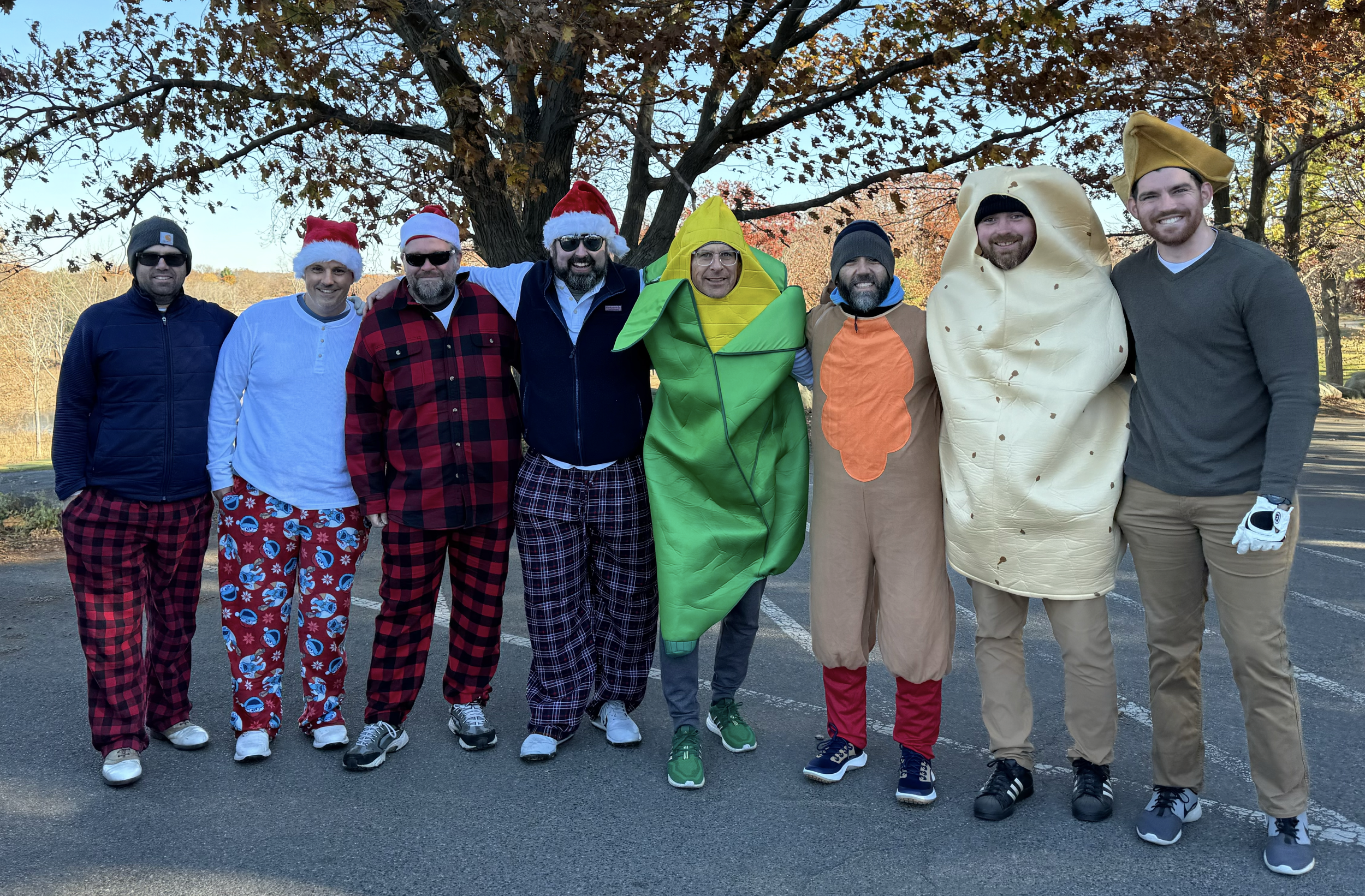 Eight men pose outdoors in festive costumes, including Santa hats, plaid pajamas, an ear of corn, a turkey, and a pie.