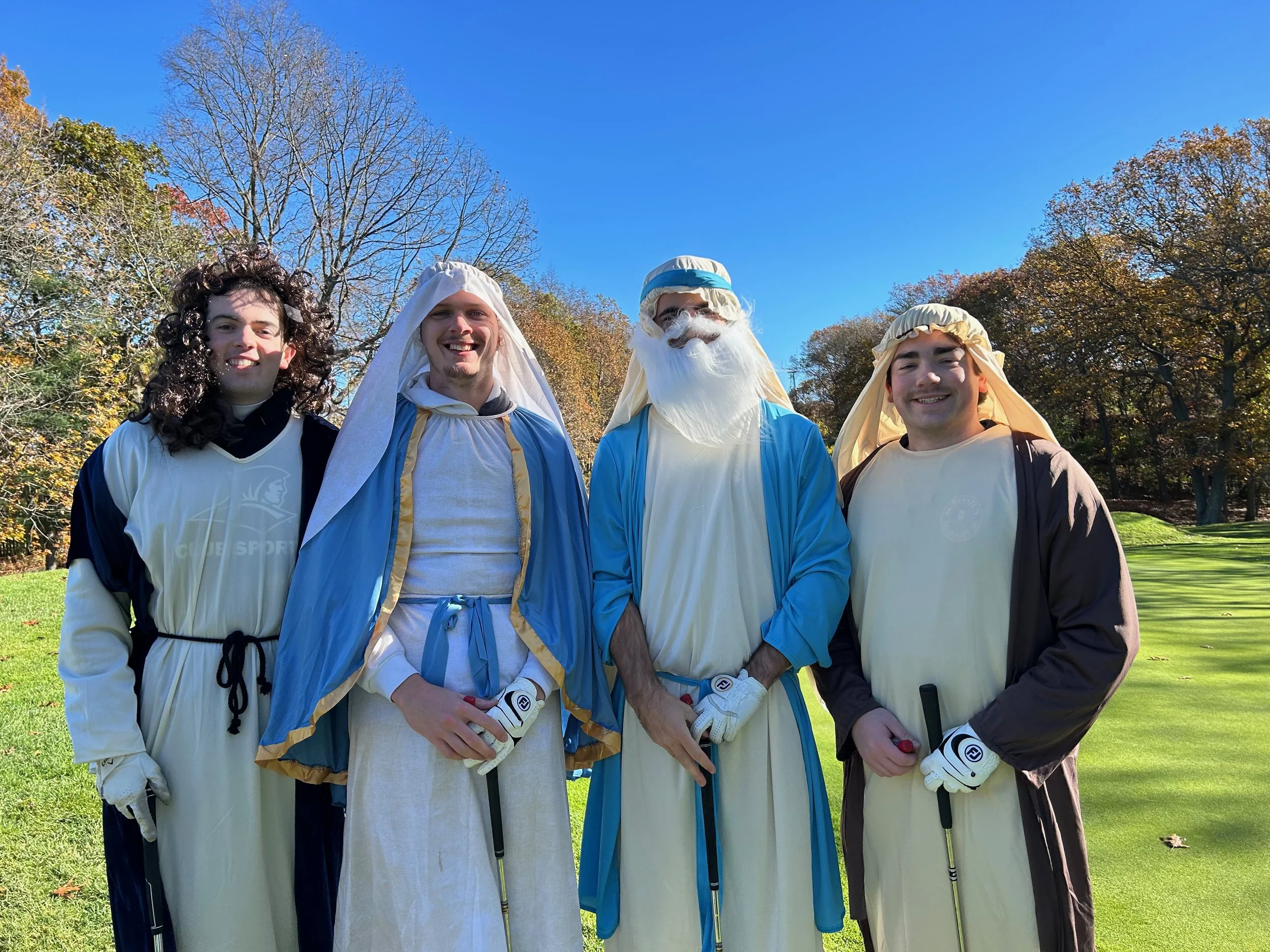 Four people in colorful biblical costumes pose with golf clubs on a sunny golf course.