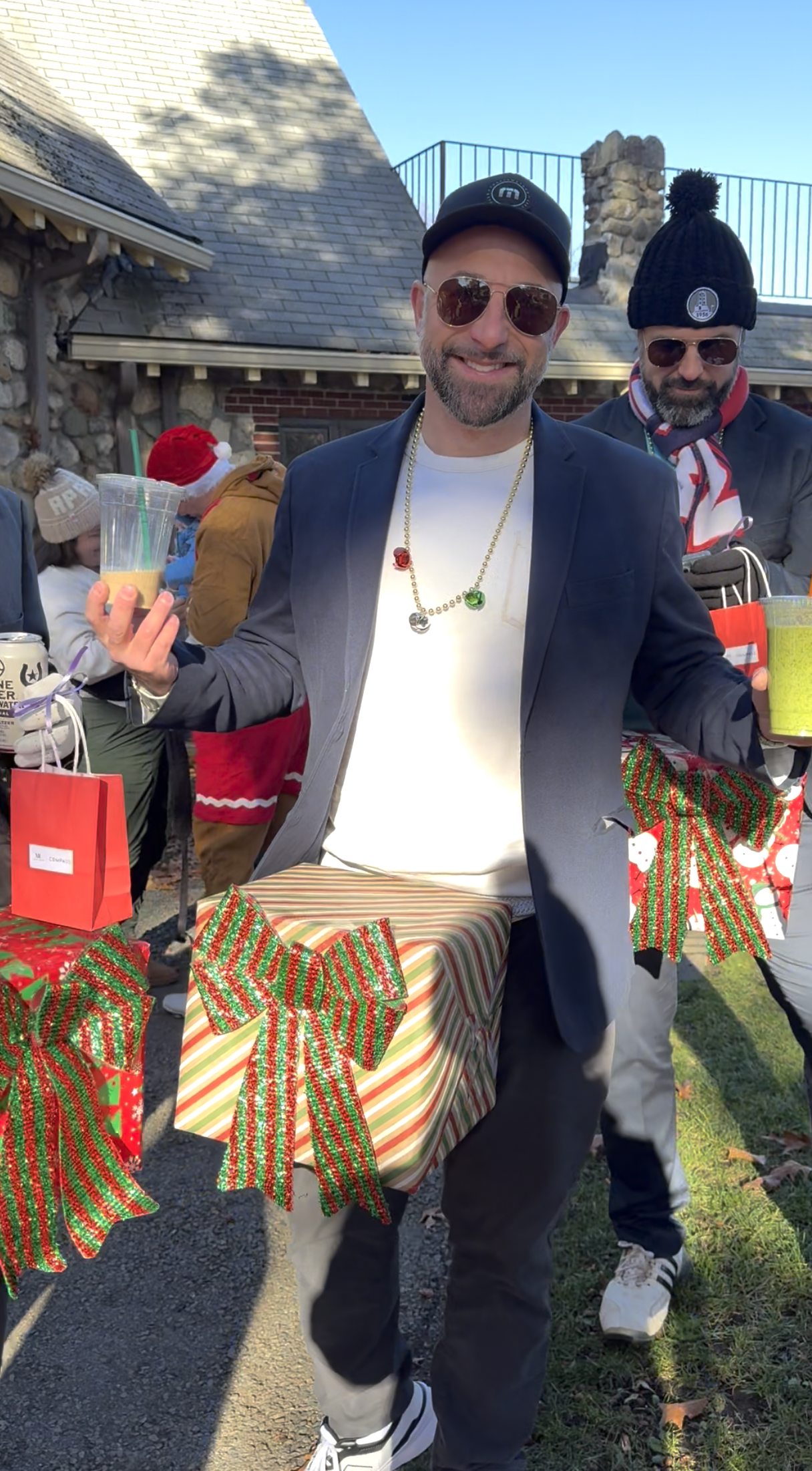 Smiling man in sunglasses and a black cap holds drinks beside wrapped Christmas presents at an outdoor holiday gathering.