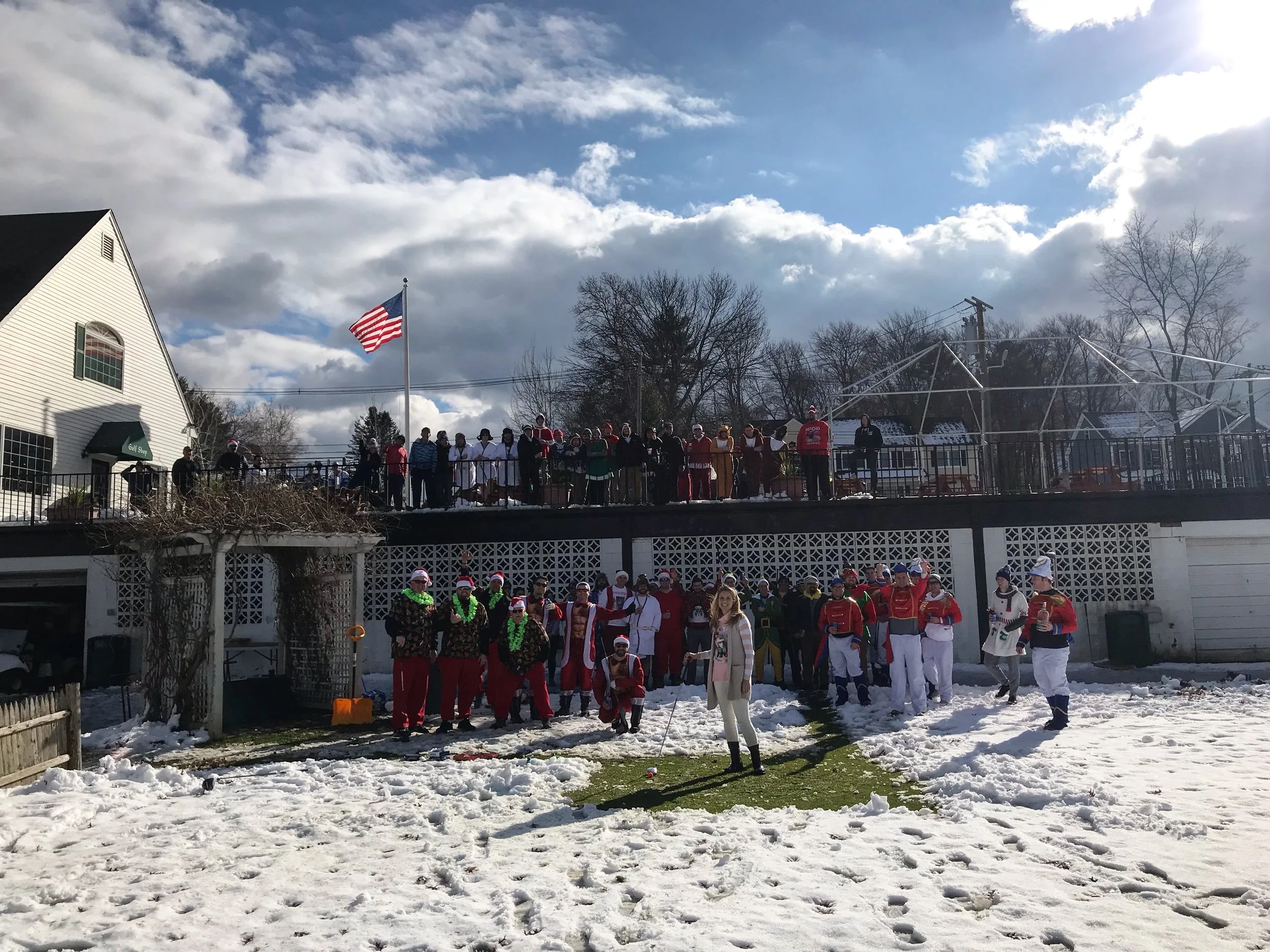 Large group in colorful holiday costumes gathered on a snowy golf course beside an American flag and clubhouse.