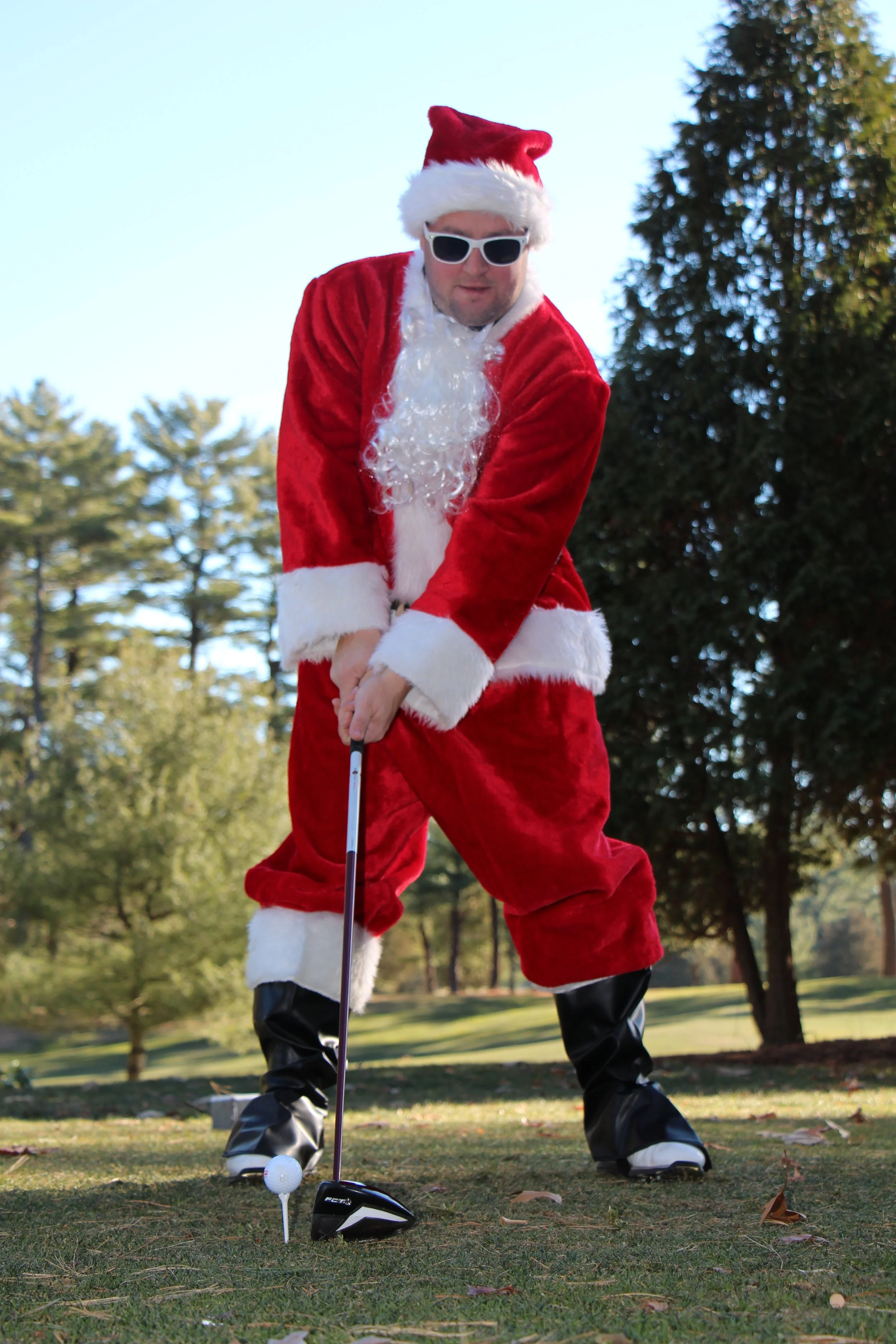 Santa Claus wearing sunglasses plays golf outdoors on a grassy course.