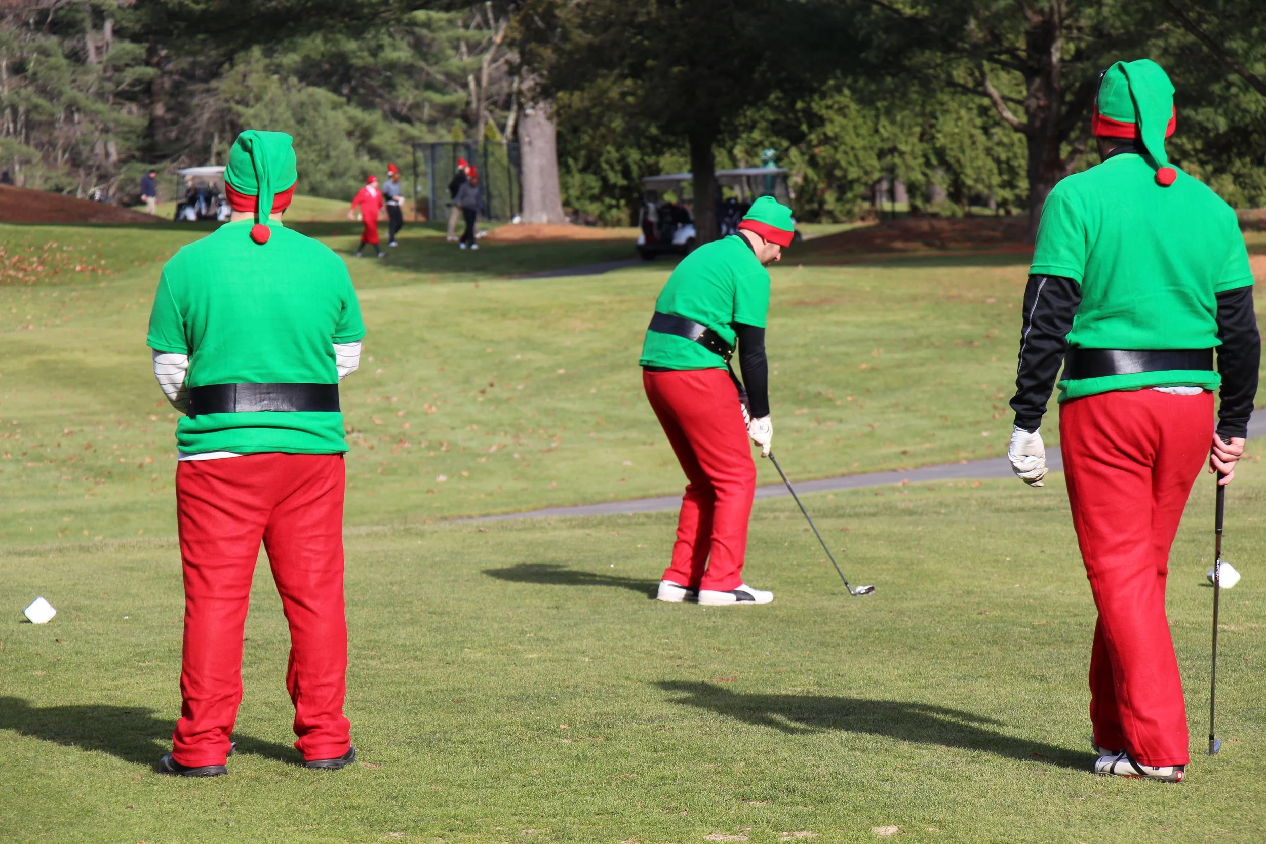 Three golfers dressed as Christmas elves on a golf course, with one preparing to swing.