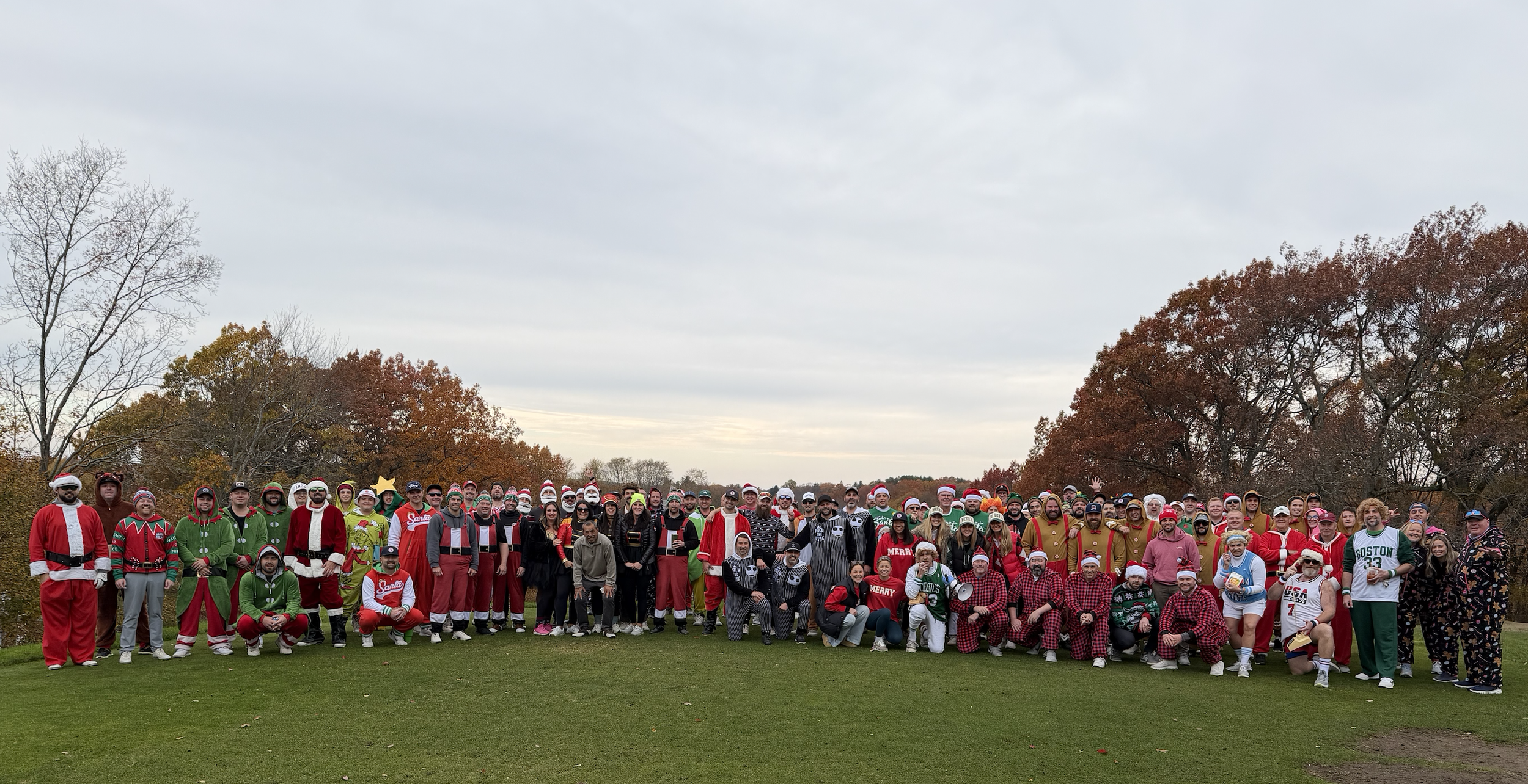 Large group wearing festive Christmas costumes and sweaters posing on a grassy field in autumn