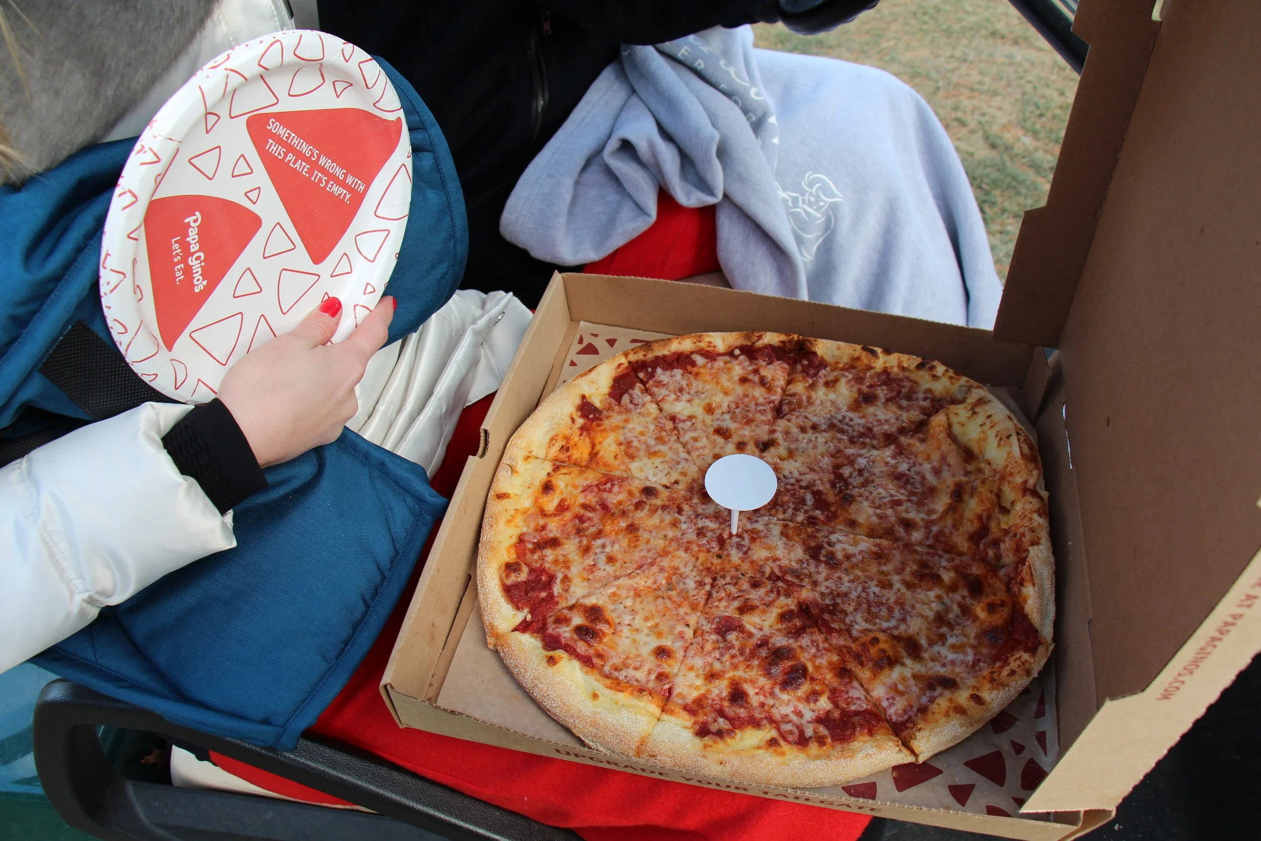 Large cheese pizza in an open box beside a hand holding a patterned paper plate and a gray hoodie.