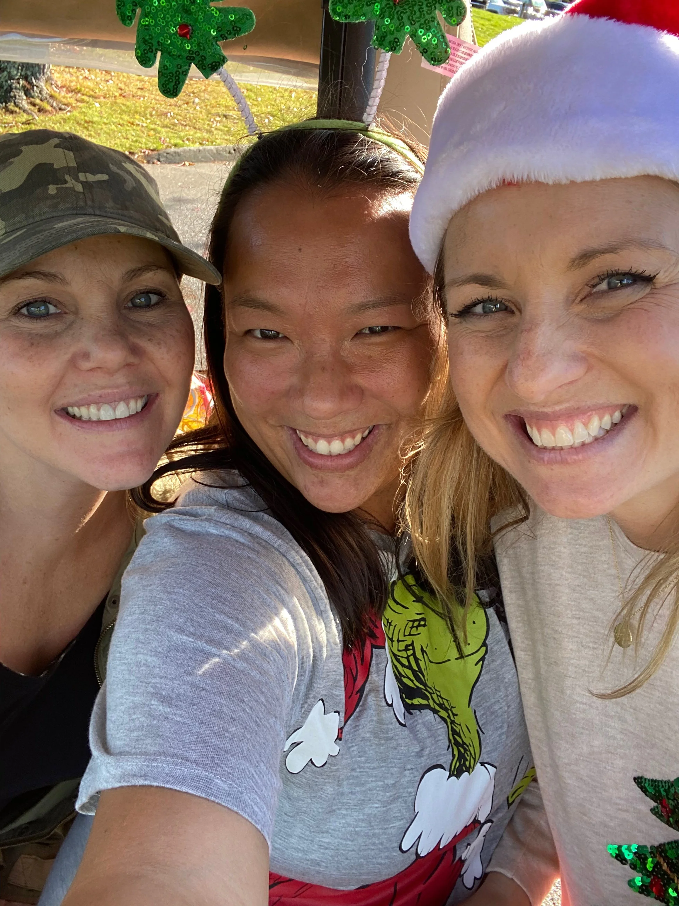 Three smiling adults take a close-up selfie outdoors, wearing festive holiday hats and colorful sweaters.