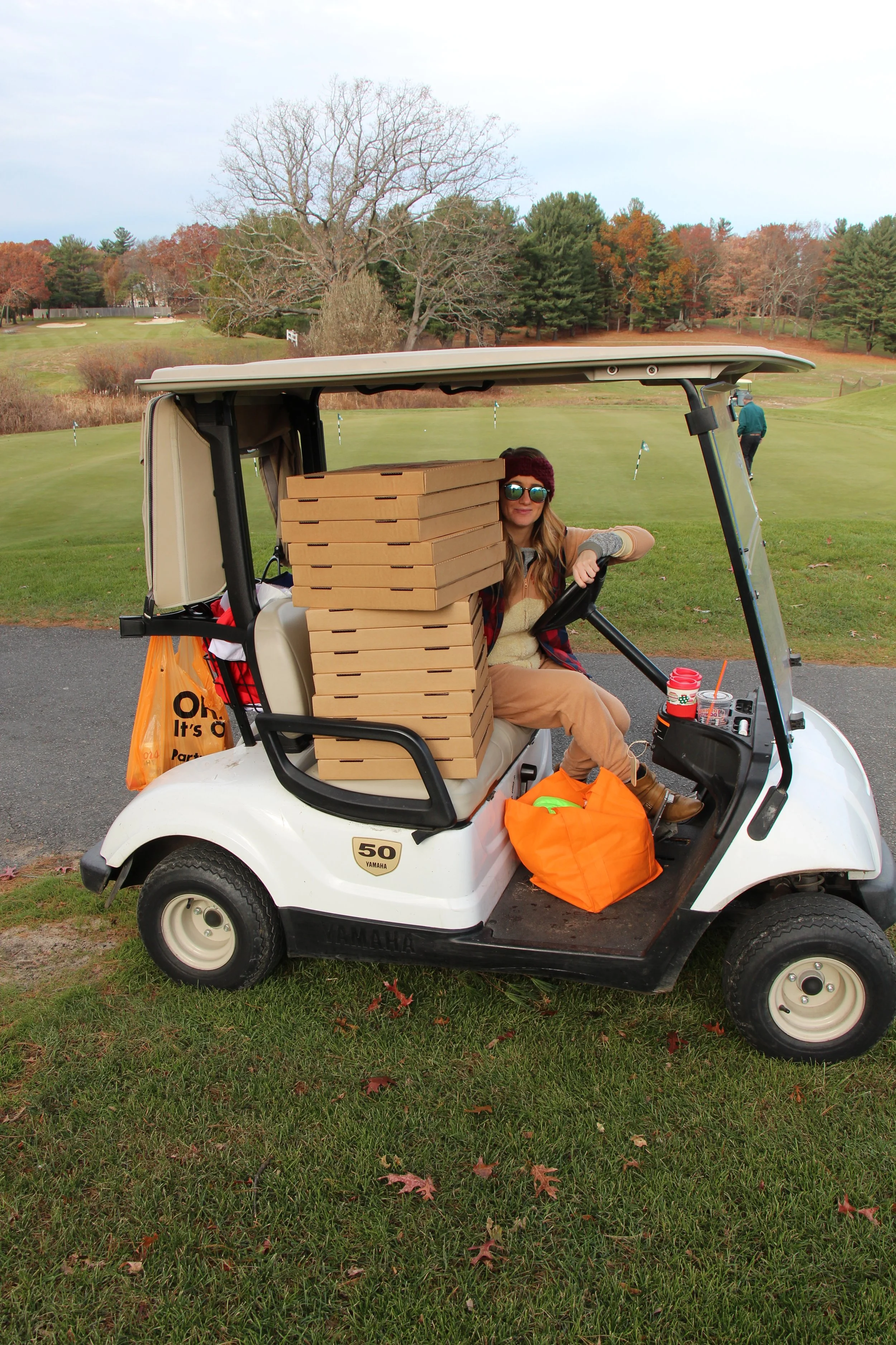 Woman sitting in a golf cart loaded with stacked pizza boxes on a golf course in autumn