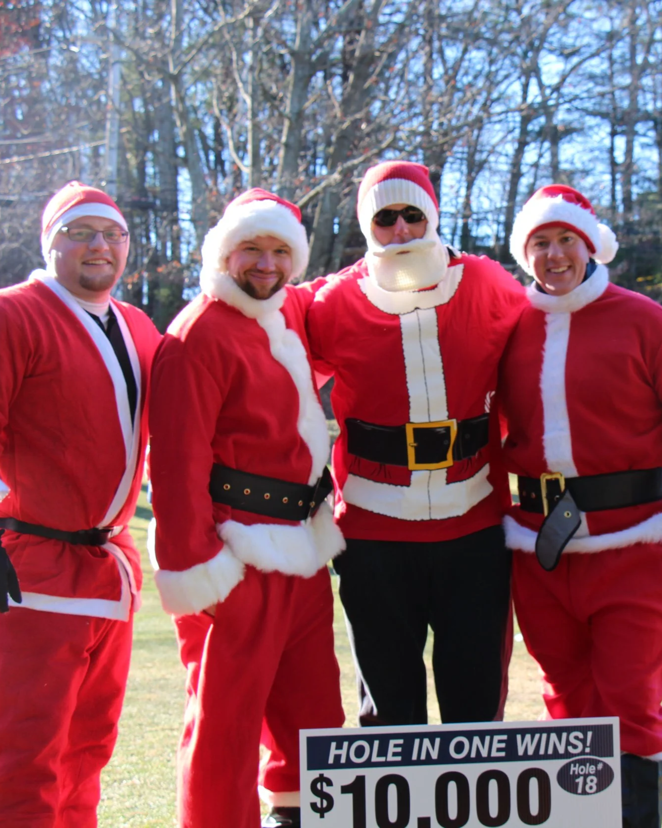 Four people dressed as Santa Claus pose outdoors behind a sign offering a $10,000 hole-in-one prize.