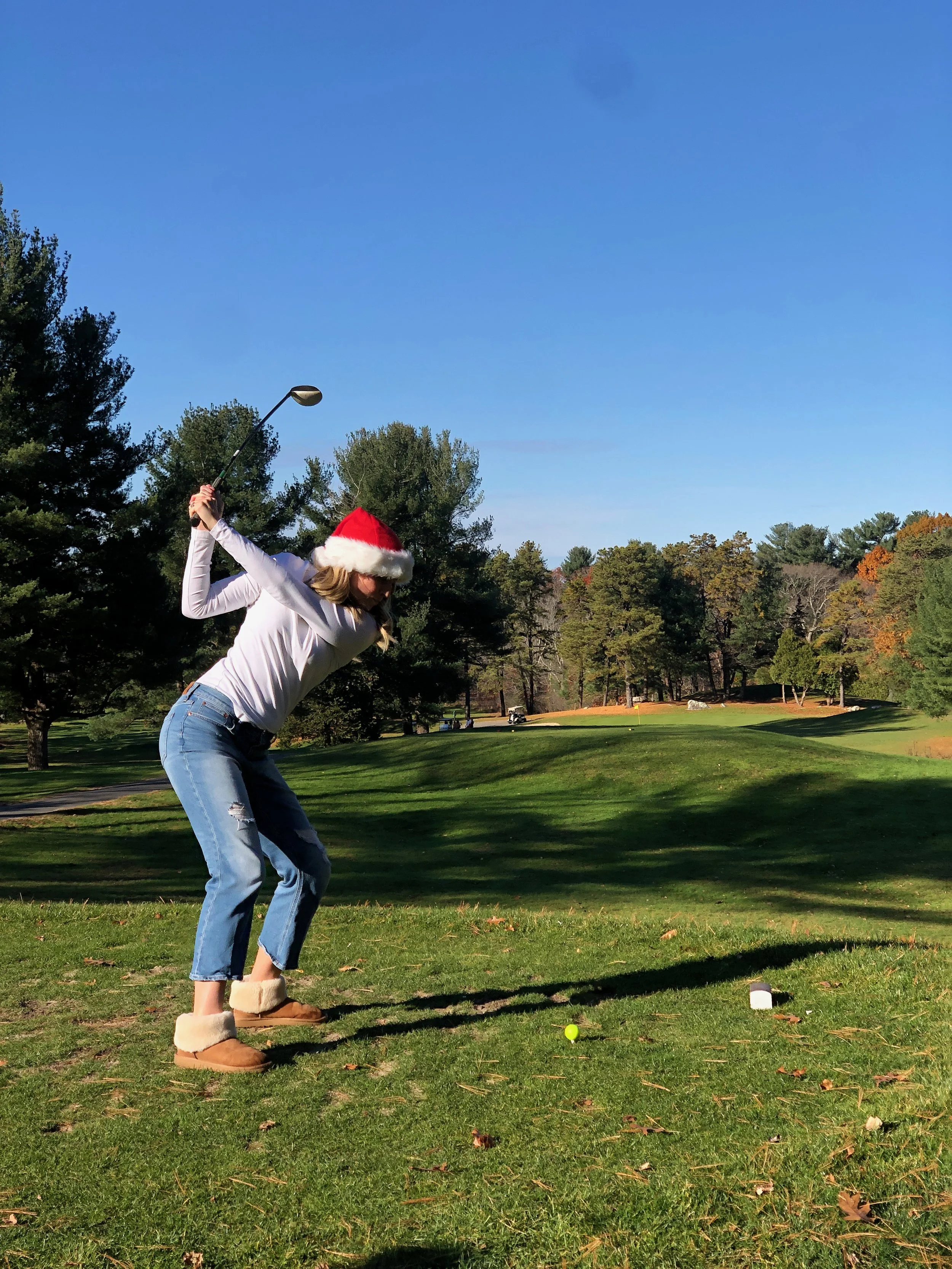 Woman wearing a Santa hat swings a golf club on a sunny golf course.