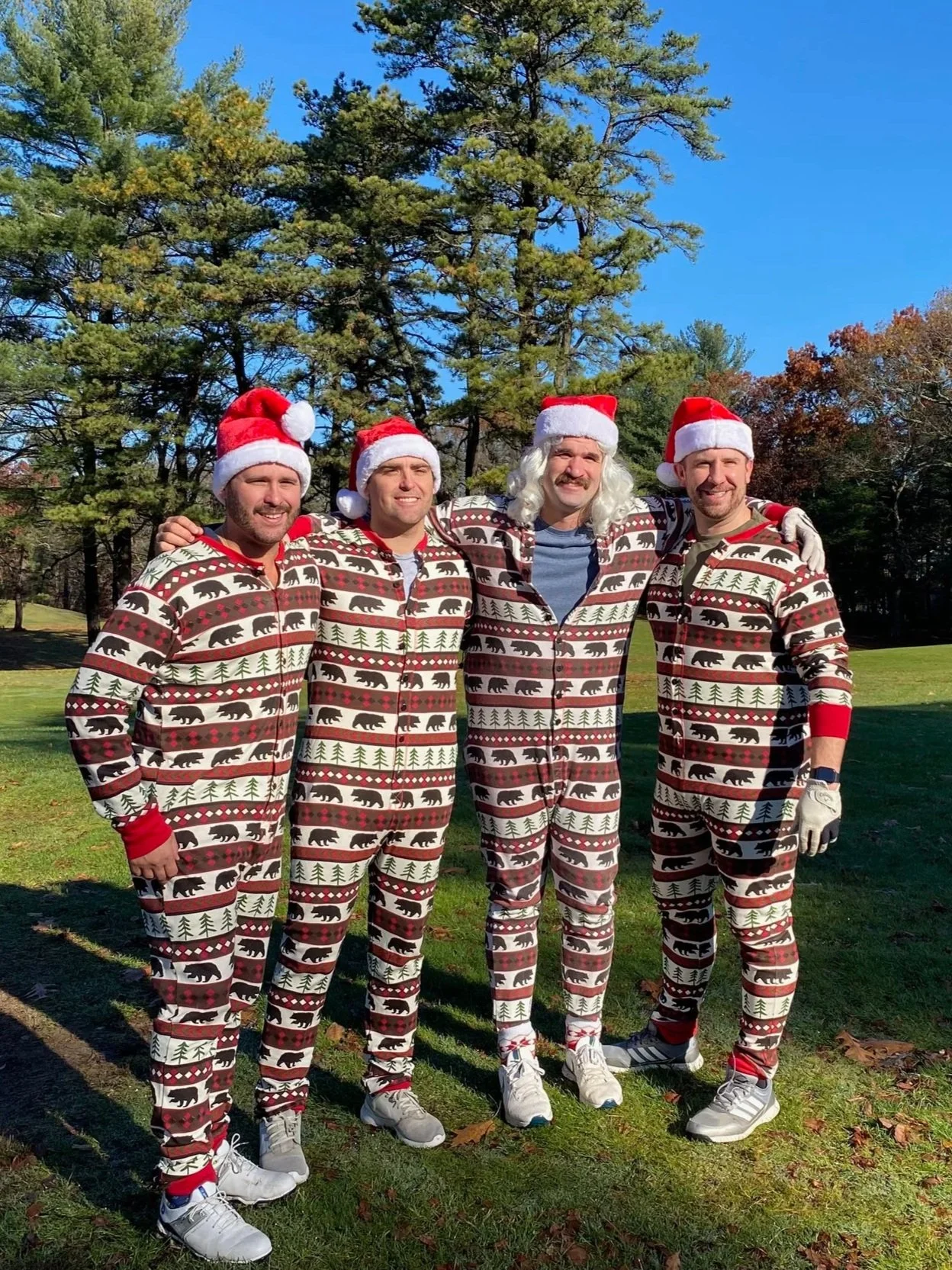 Four men wearing Santa hats and matching bear-patterned Christmas pajamas pose outdoors in a park.