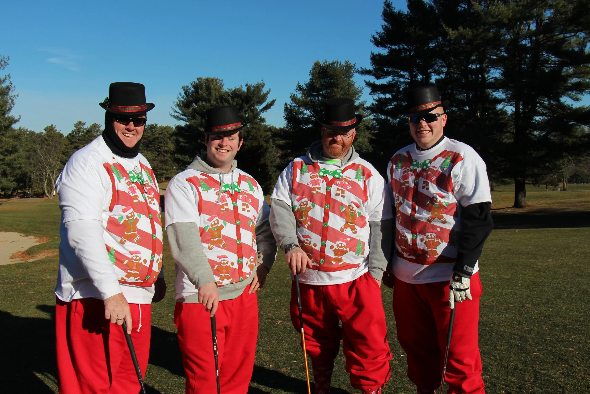 Four men in festive Christmas-themed outfits and top hats posing with golf clubs in a park