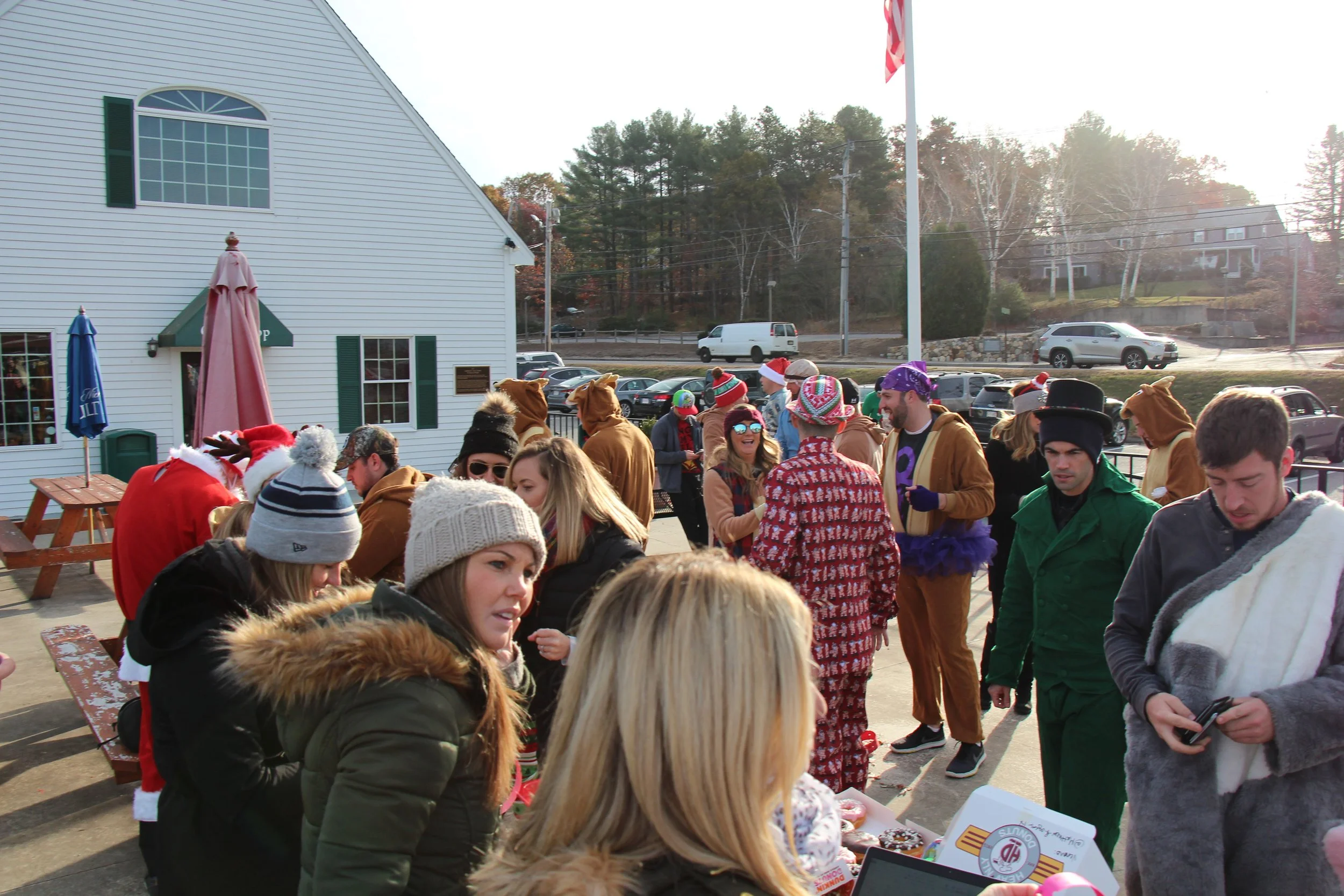 Crowd in festive Christmas costumes gathered outside a white building with picnic tables and an American flag