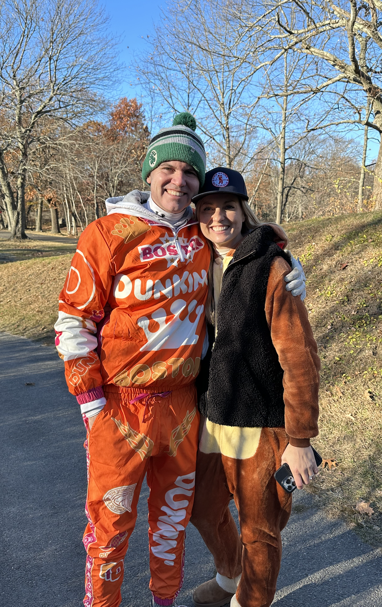 Smiling couple posing on a wooded path, with the man wearing an orange Dunkin' promotional outfit and green pom-pom beanie.