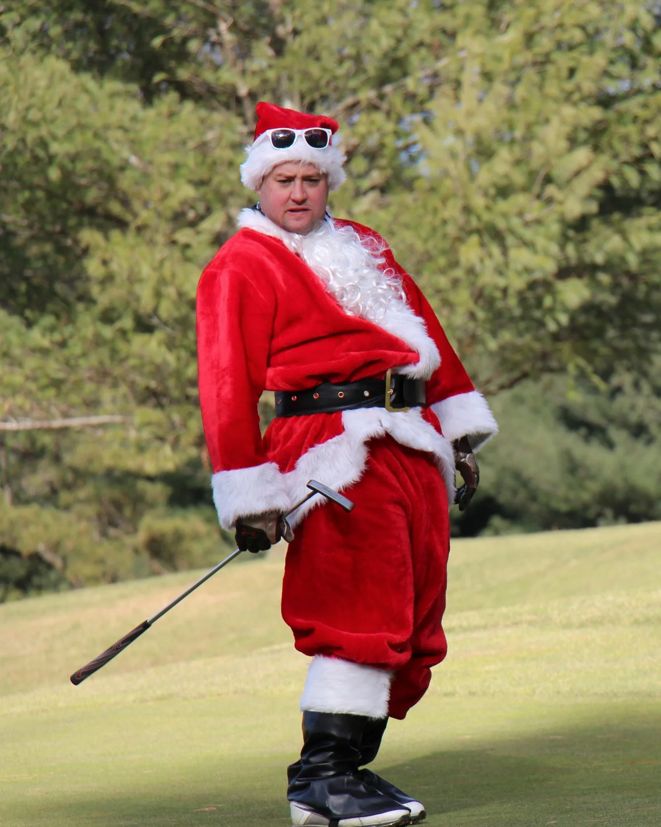 Person dressed as Santa Claus wearing sunglasses and holding a golf club on a golf course