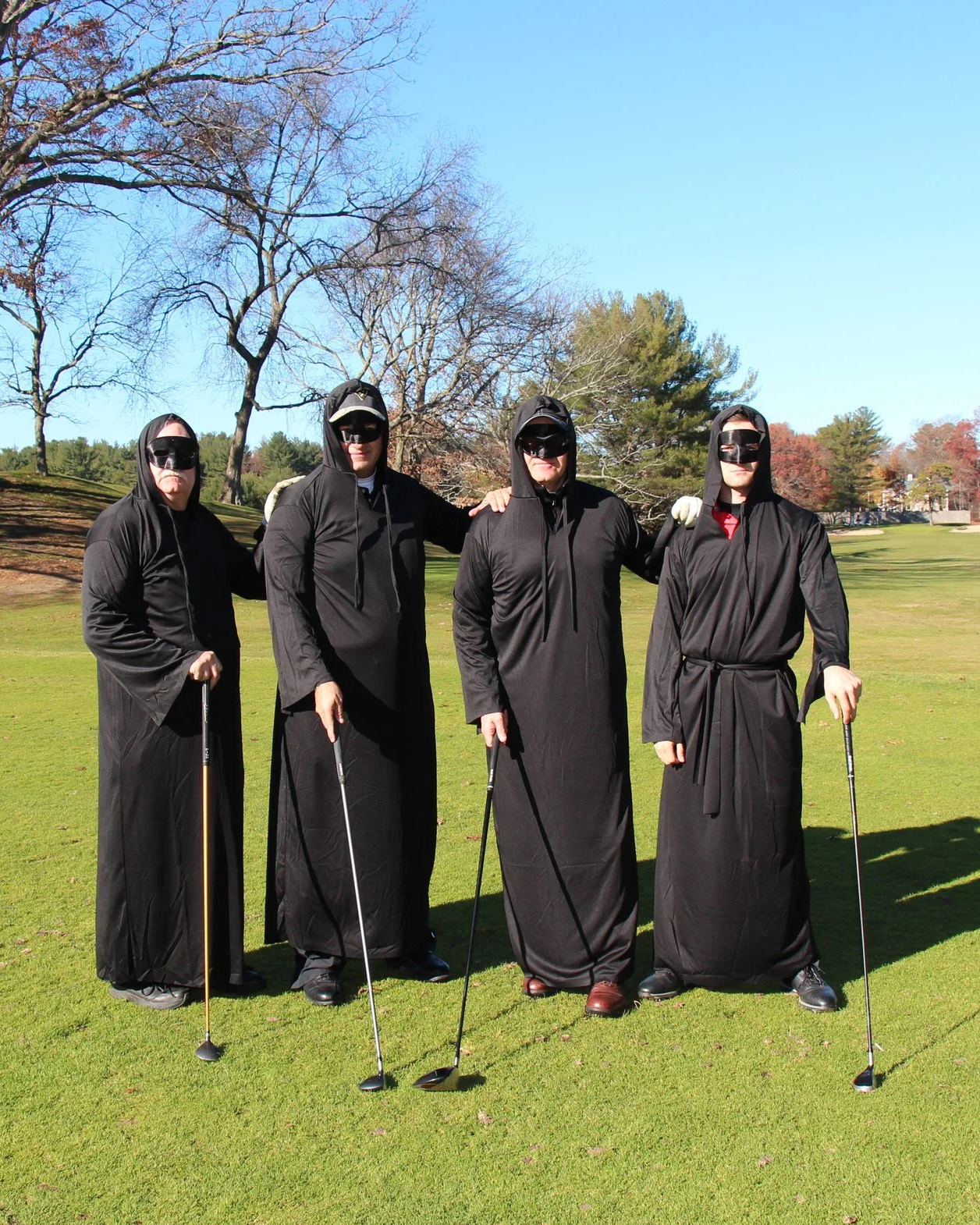 Four people dressed in black hooded costumes and masks pose with golf clubs on a sunny golf course.