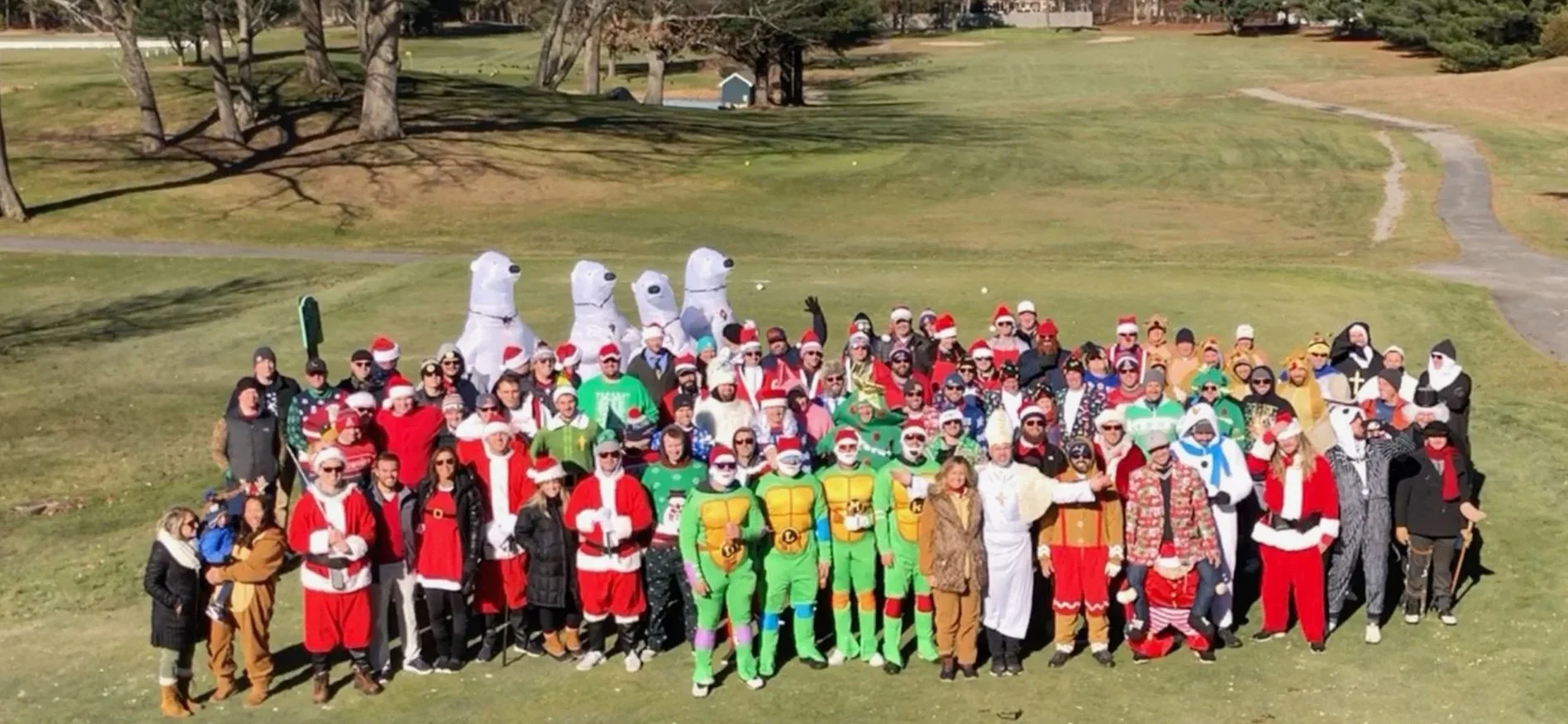 Large group in festive Christmas costumes posing on a golf course, with Santa suits, ugly sweaters, and inflatable snowmen