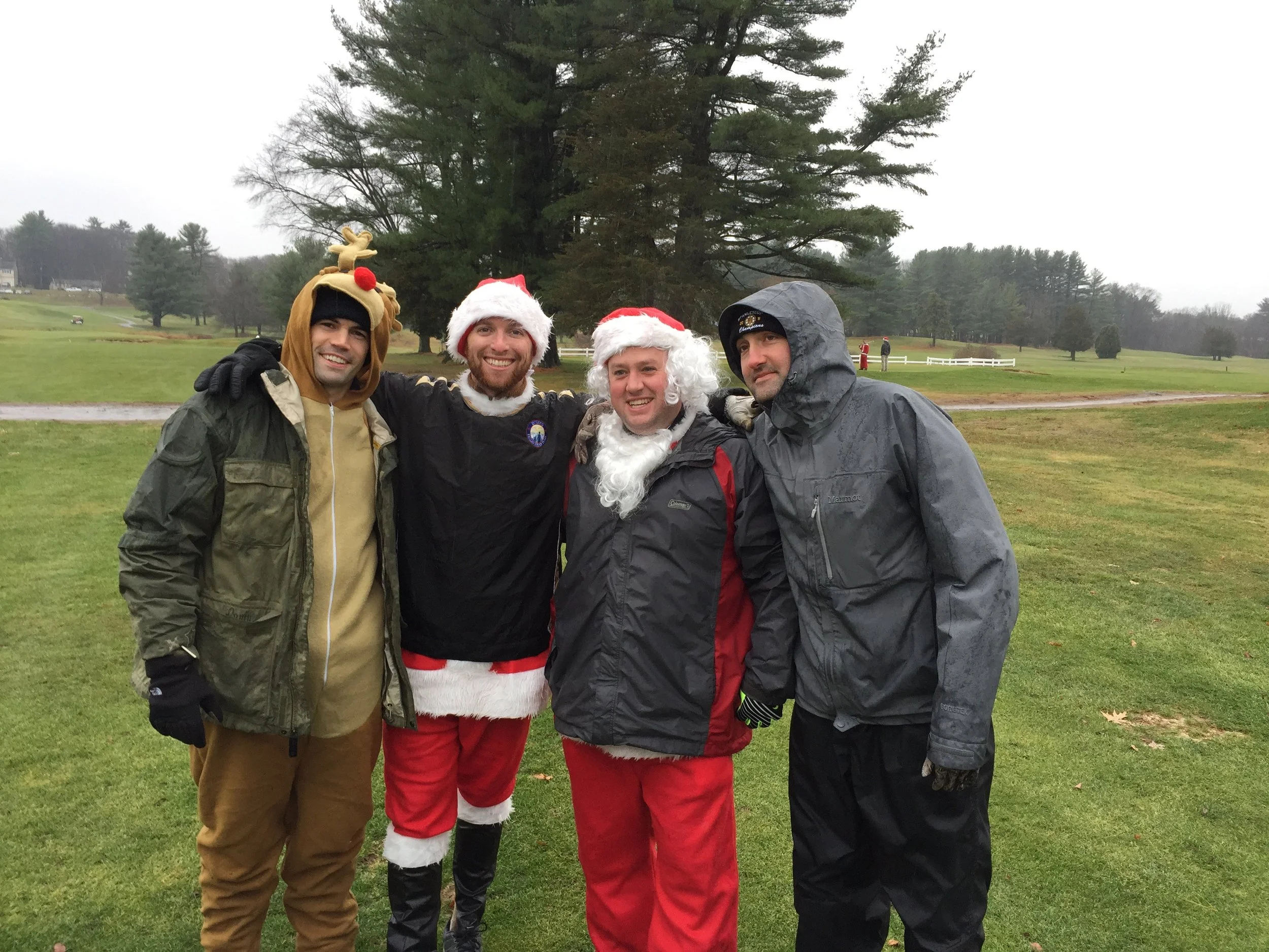 Four men in reindeer and Santa costumes pose outdoors on a rainy golf course.