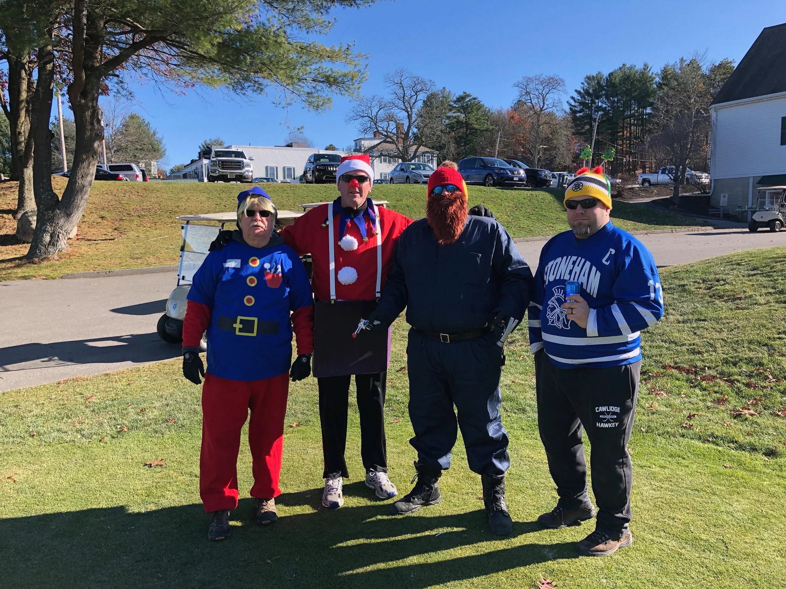Four adults in festive costumes pose outdoors on a sunny day, including Santa Claus, a snowman, a bearded winter character, and a hockey fan.