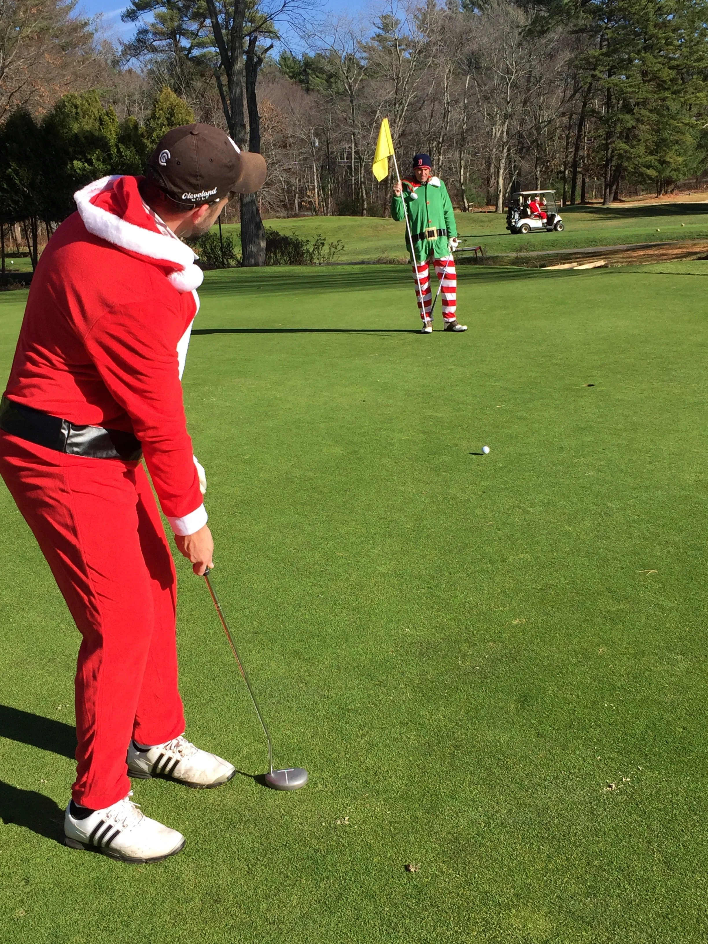 Golfer in a Santa suit putting on a golf green while another golfer dressed as an elf holds a yellow flag.