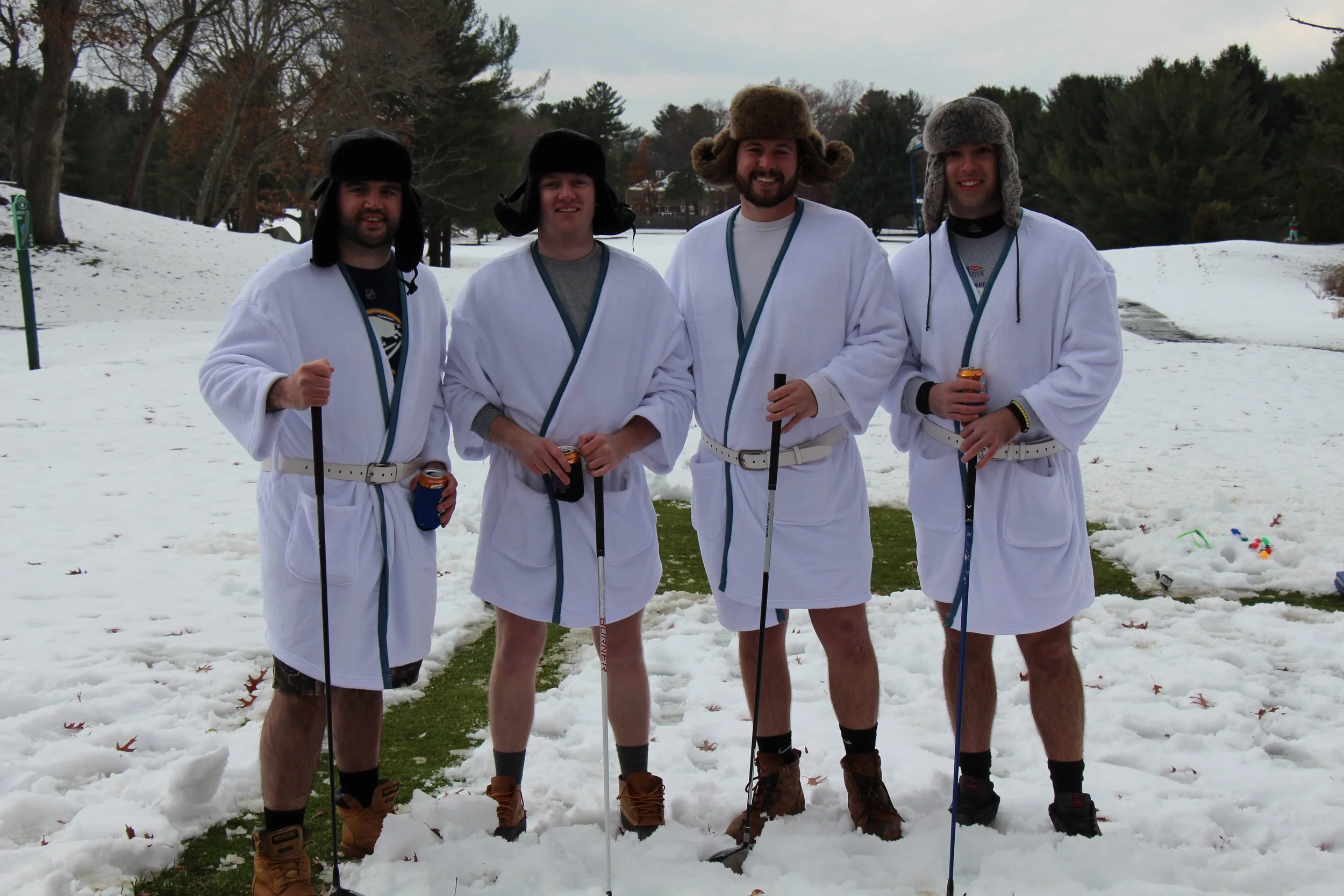 Four men in white robes and winter hats pose with ski poles in a snowy park.