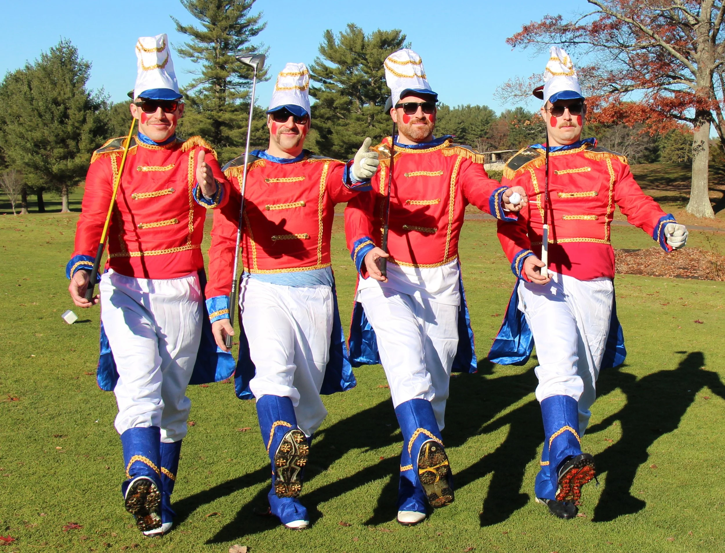 Four men in red, white, and blue marching band uniforms pose with golf clubs on a sunny golf course.