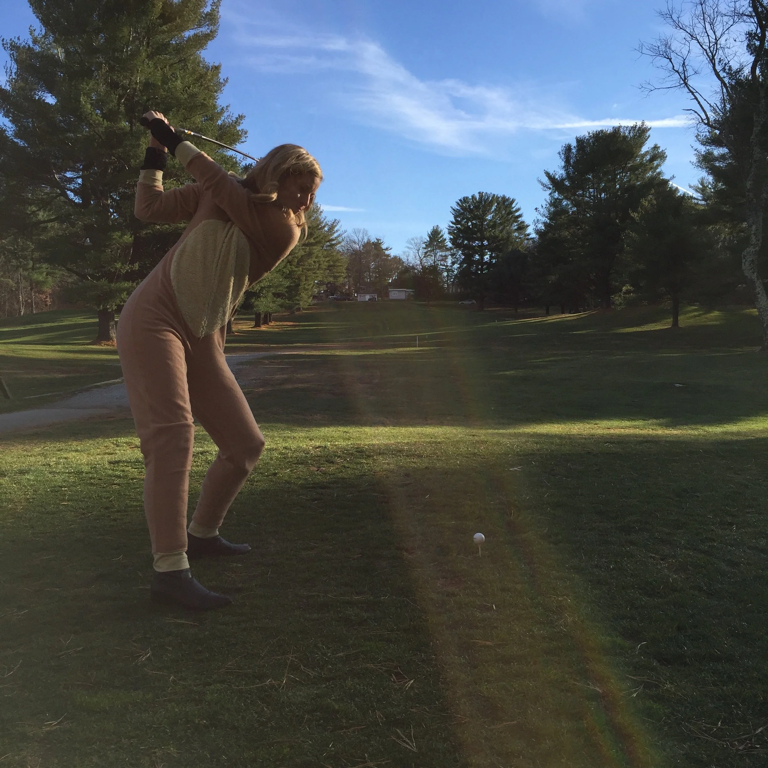Woman swinging a golf club at a golf ball on a grassy lawn surrounded by trees