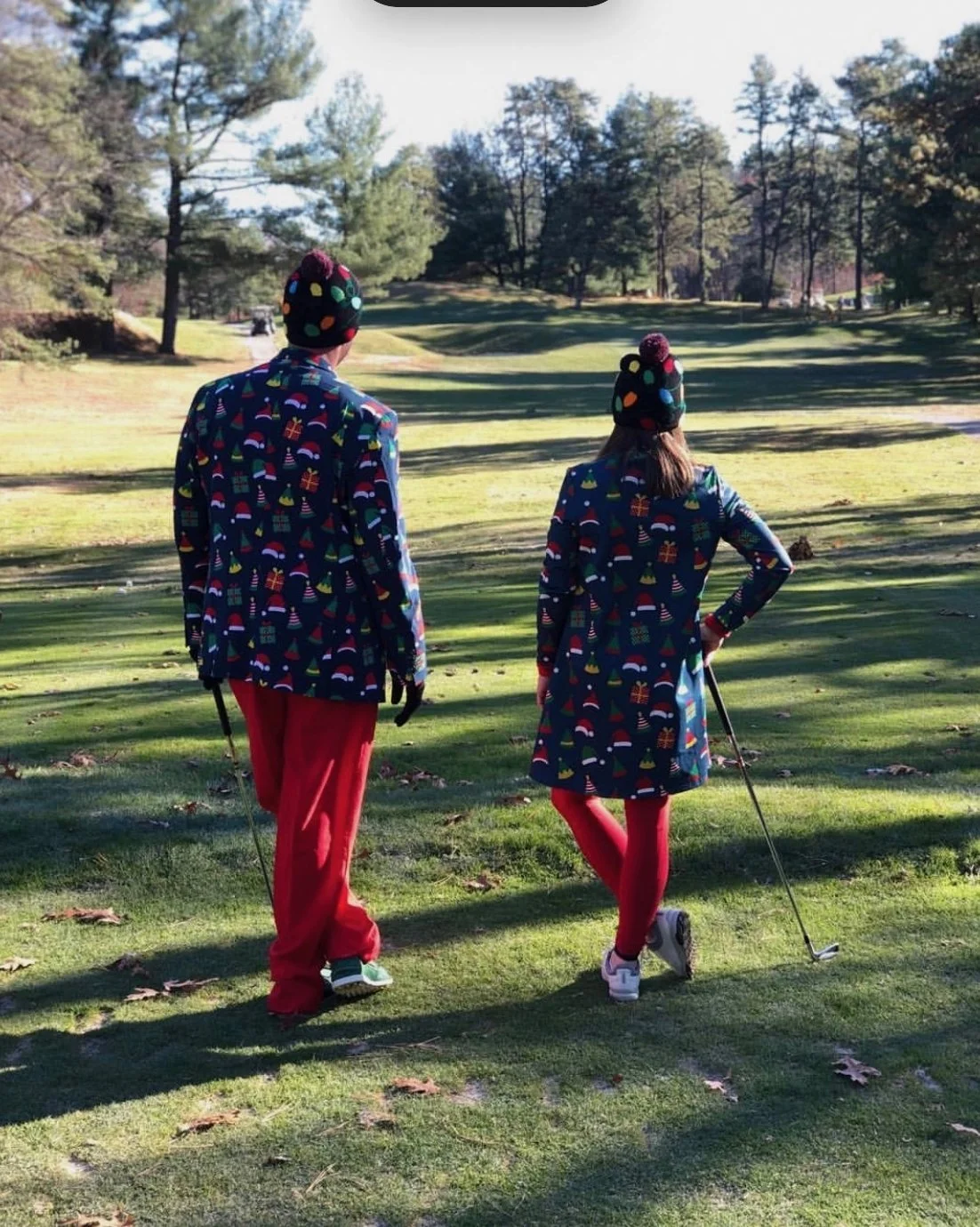 Two people in festive Christmas-patterned outfits and hats walk across a golf course carrying golf clubs.