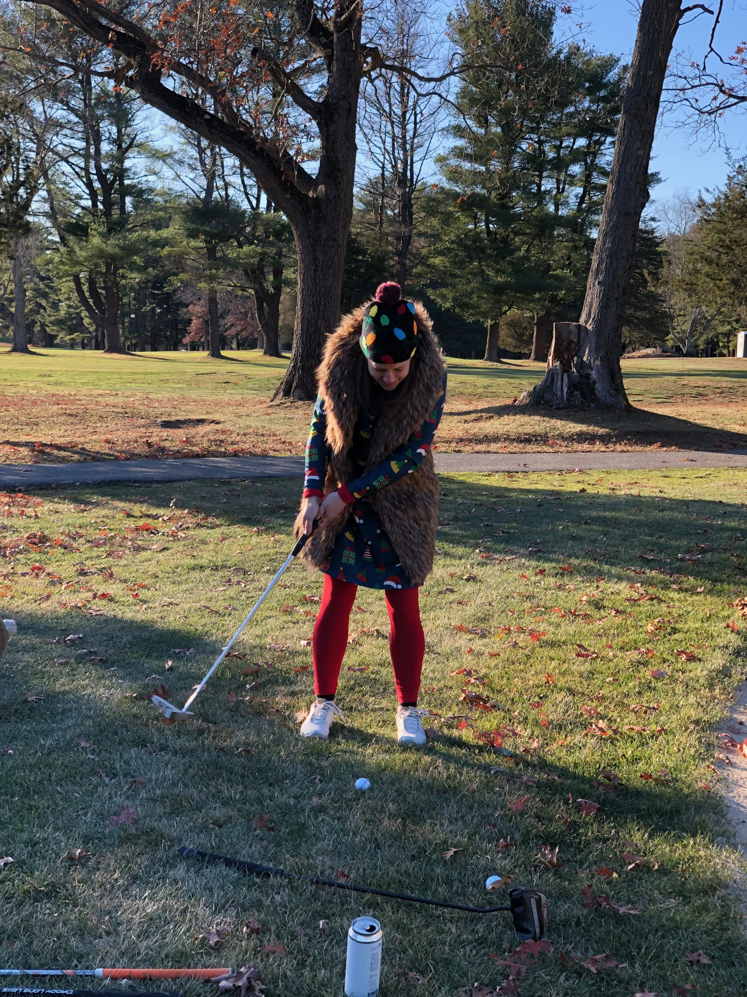 Woman in a colorful winter outfit and fur-trimmed vest holding a golf club on a tree-lined golf course.