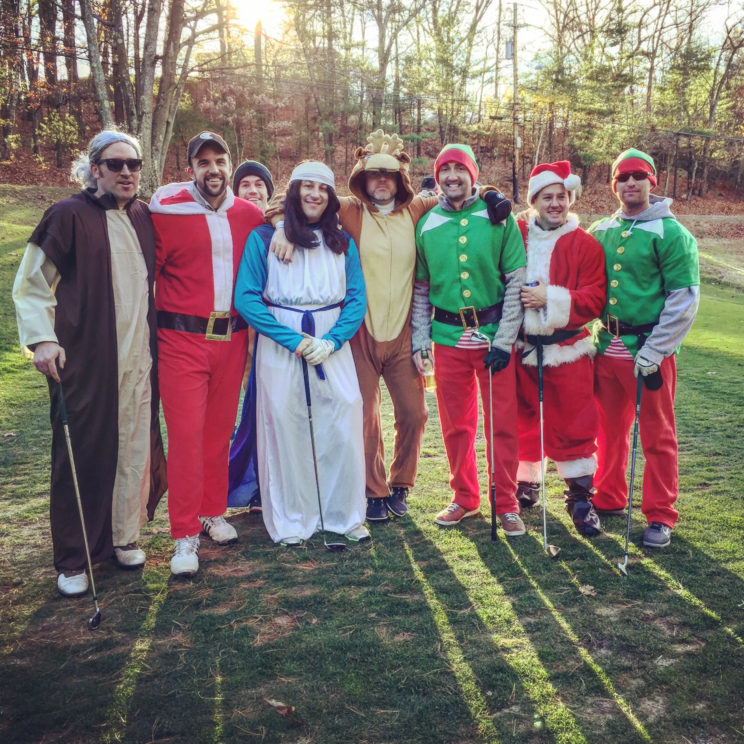 Group of people in festive Santa Claus, elf, and other holiday costumes posing outdoors in a wooded park.