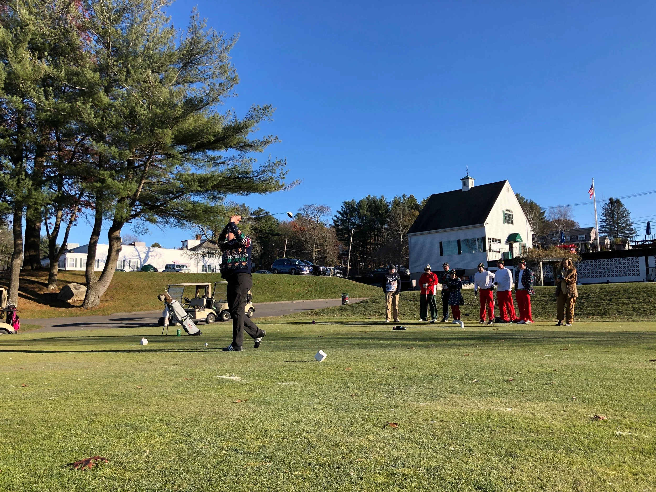 People in festive clothing playing golf on a sunny course beside a white building and American flag