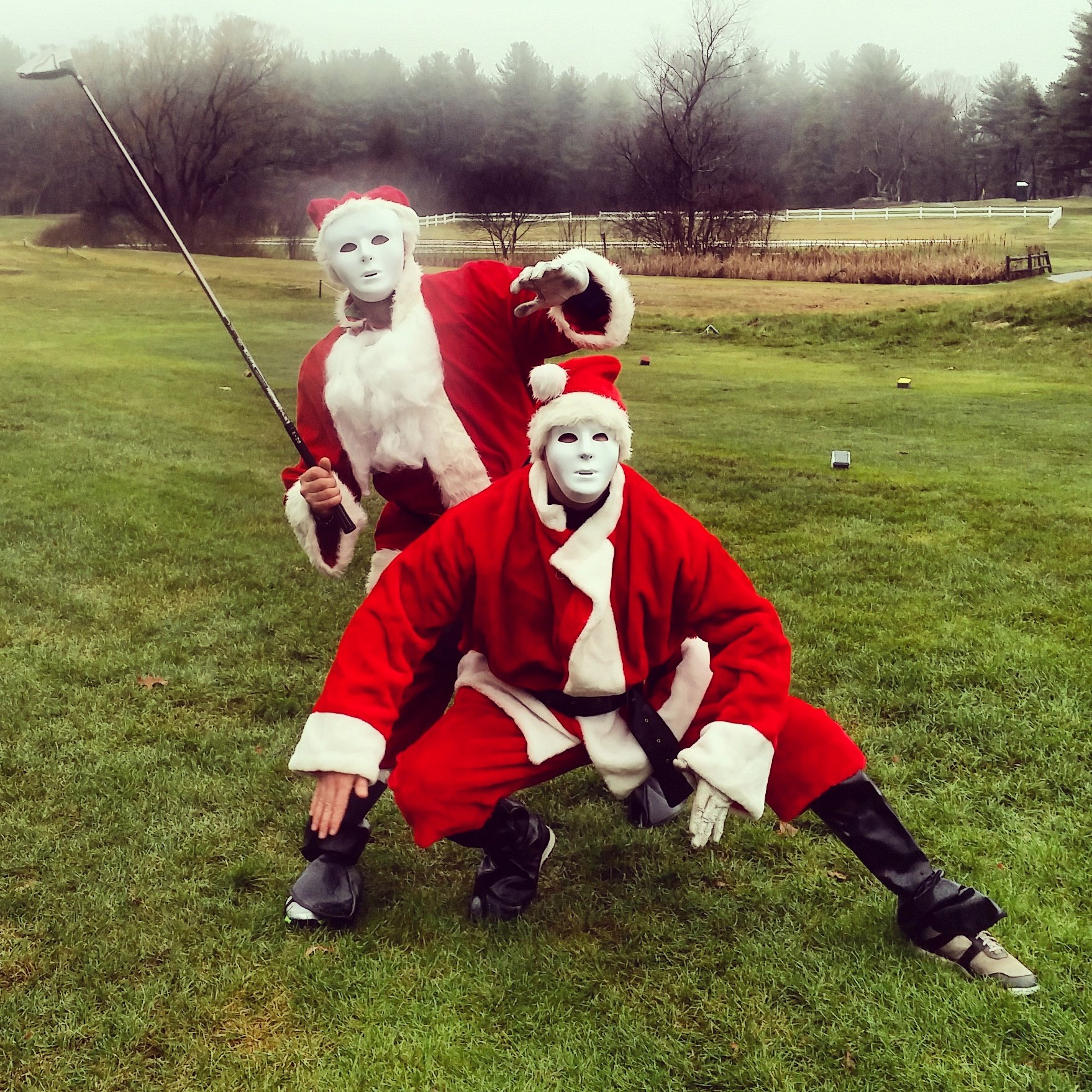 Two people in red Santa costumes and white masks pose on a grassy field, with one holding a golf club.