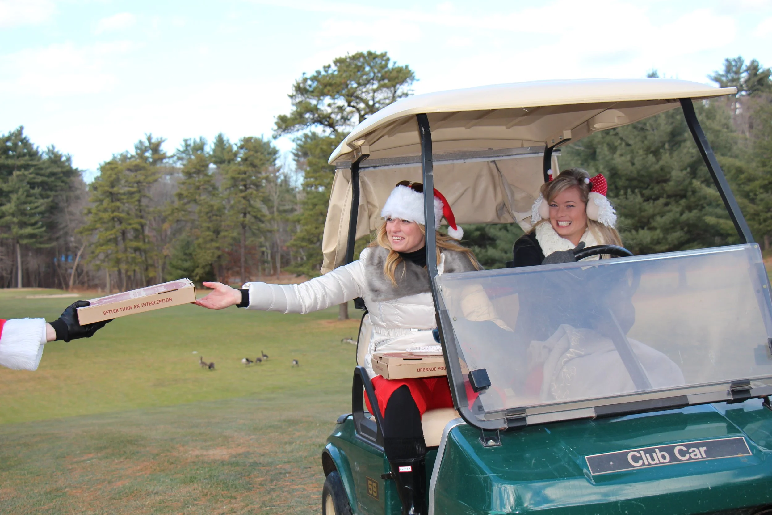 Two women in festive winter clothing ride a golf cart while Santa hands them a pizza box on a golf course.