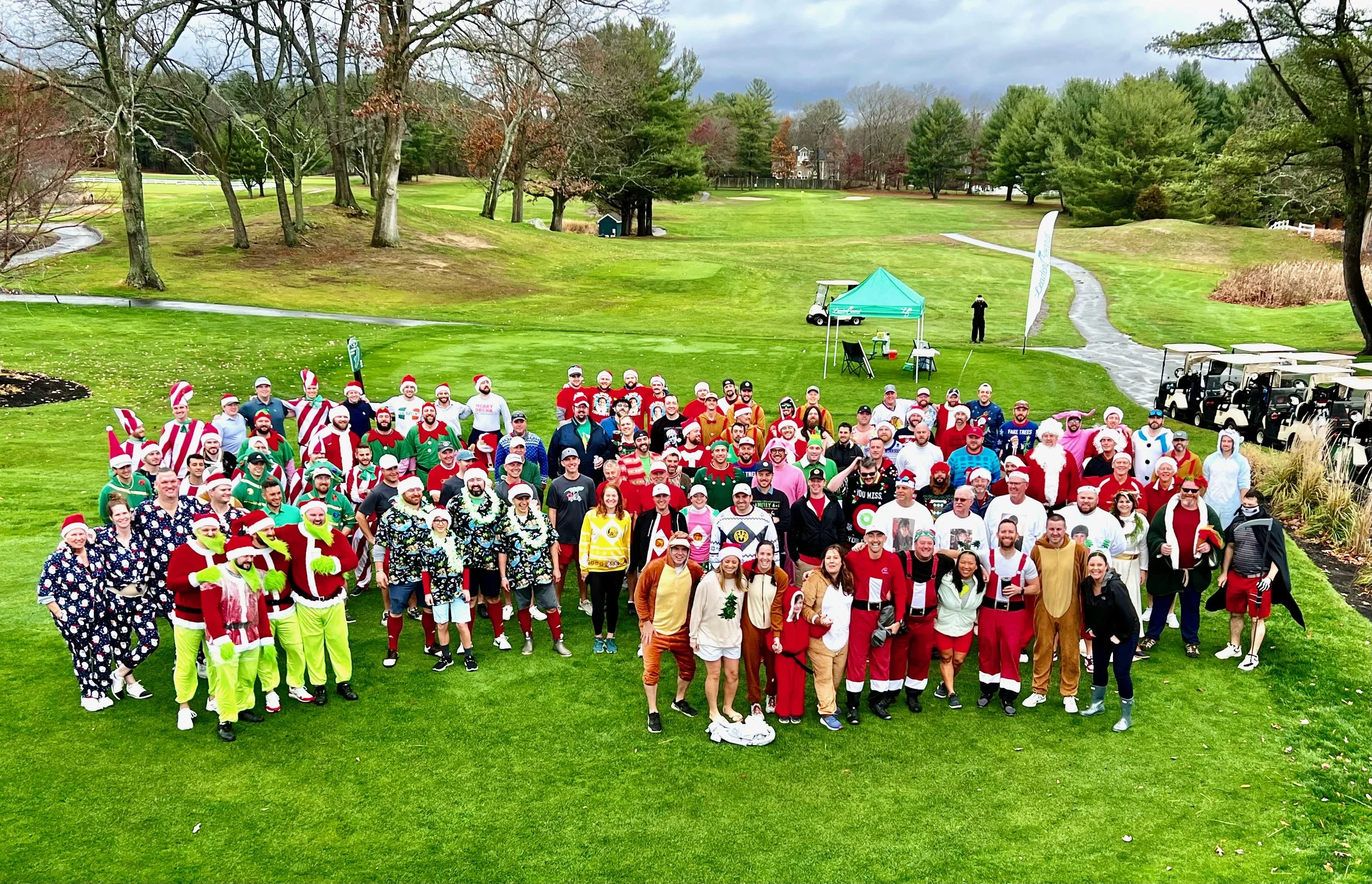 Large group in festive Christmas costumes posing on a golf course with golf carts and trees in the background