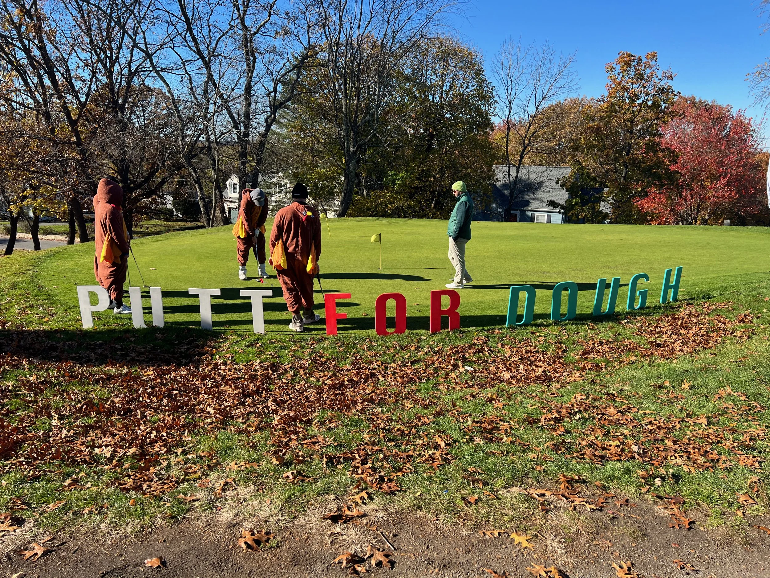 Golfers on an autumn putting green beside large colorful letters spelling “PUTT FOR DOUGH.”