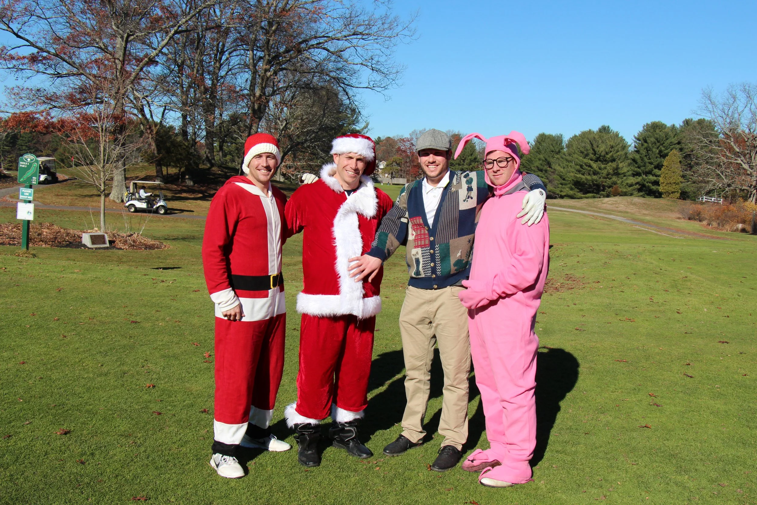 Four people in Santa, elf, and pink bunny costumes pose on a golf course beside a golf cart.