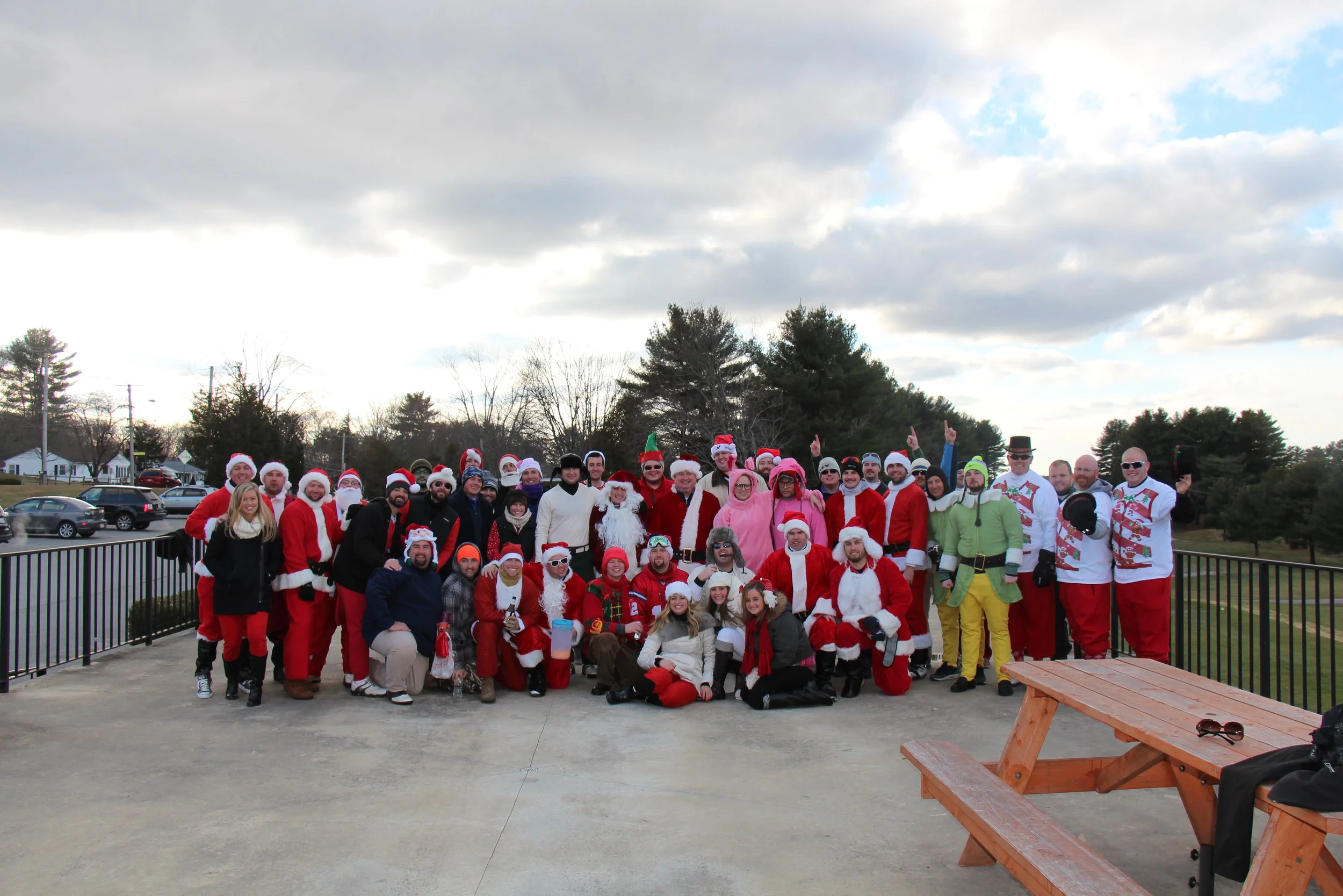 Large group dressed in Santa Claus and other festive costumes posing outdoors on a patio beneath a cloudy sky.