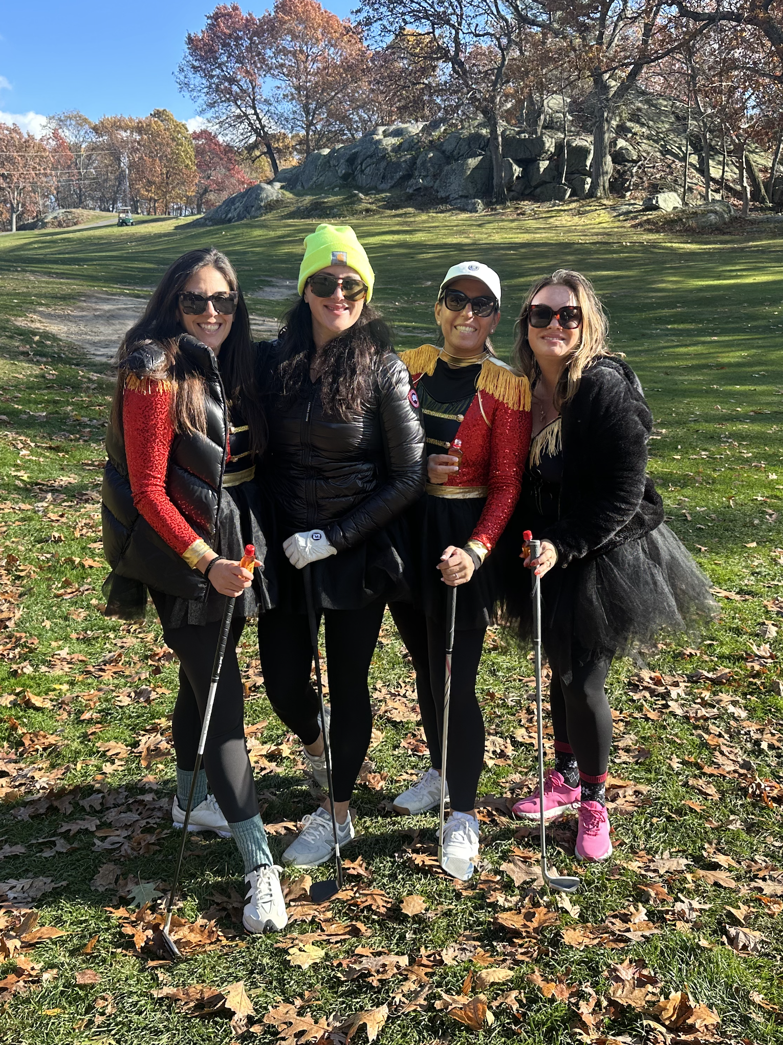 Four women pose outdoors on a sunny autumn day, wearing costumes and holding golf clubs in front of rocky terrain and colorful trees.